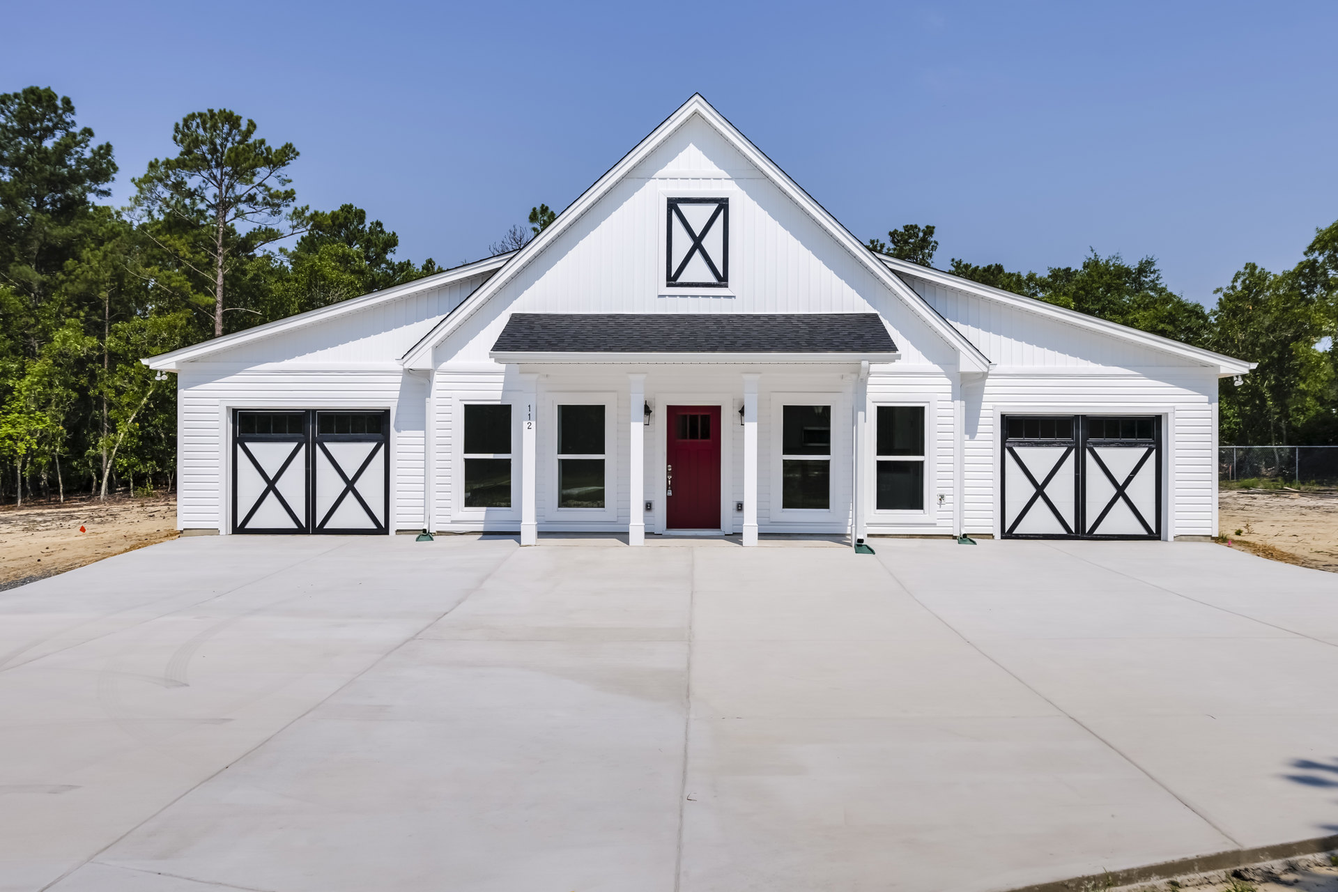 White siding house with a bold red front door, black and white address sign, white garage door, shaded porch, and surrounding trees.
