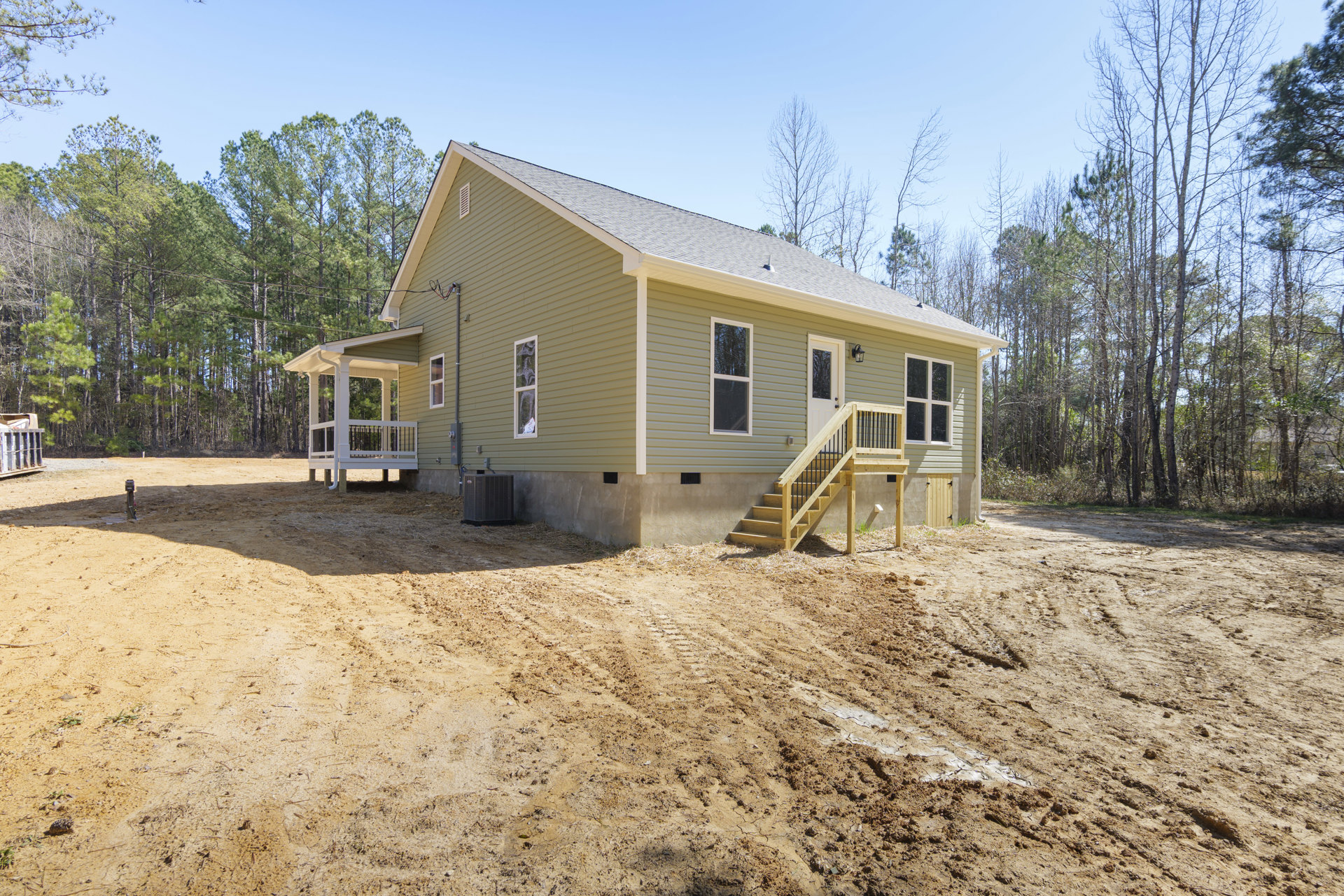 Two-story home with white-framed windows, wooden porch stairs, and bare dirt yard; metal container near entry and leafy trees in background