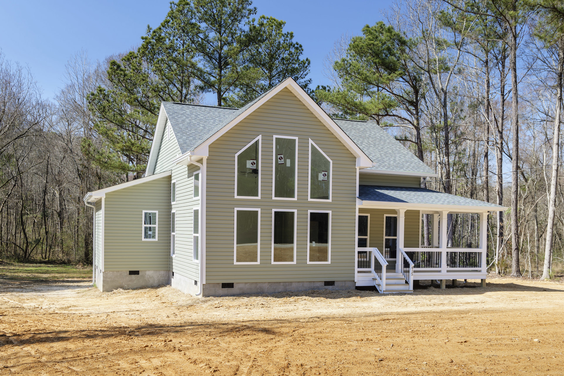 Two-story house with multiple white-framed windows, covered front porch, dirt yard, and trees in the background; sign visible in one window.