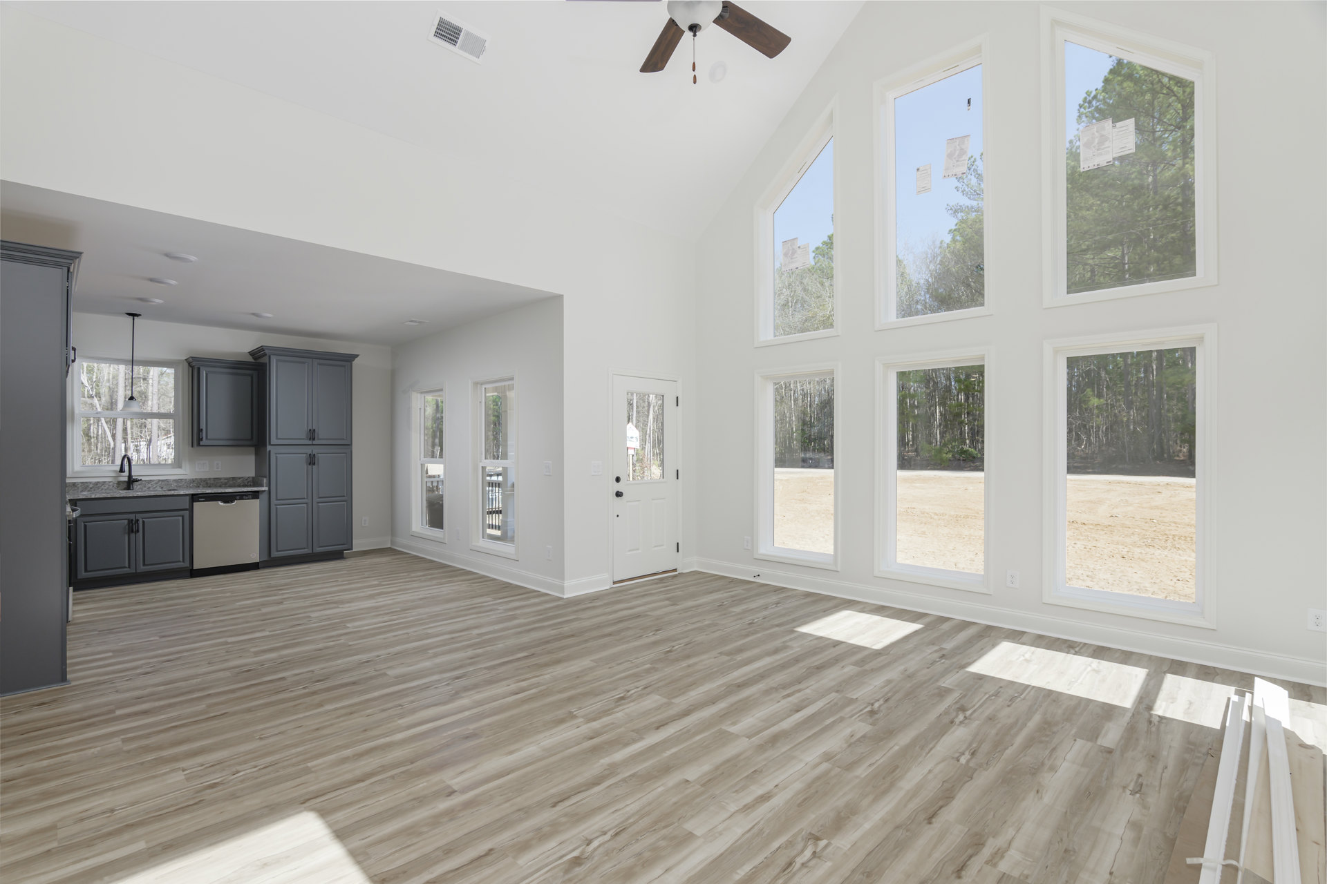 Spacious room with wood flooring, grey cabinetry with black handles, ceiling fan, multiple windows, and white wall boards