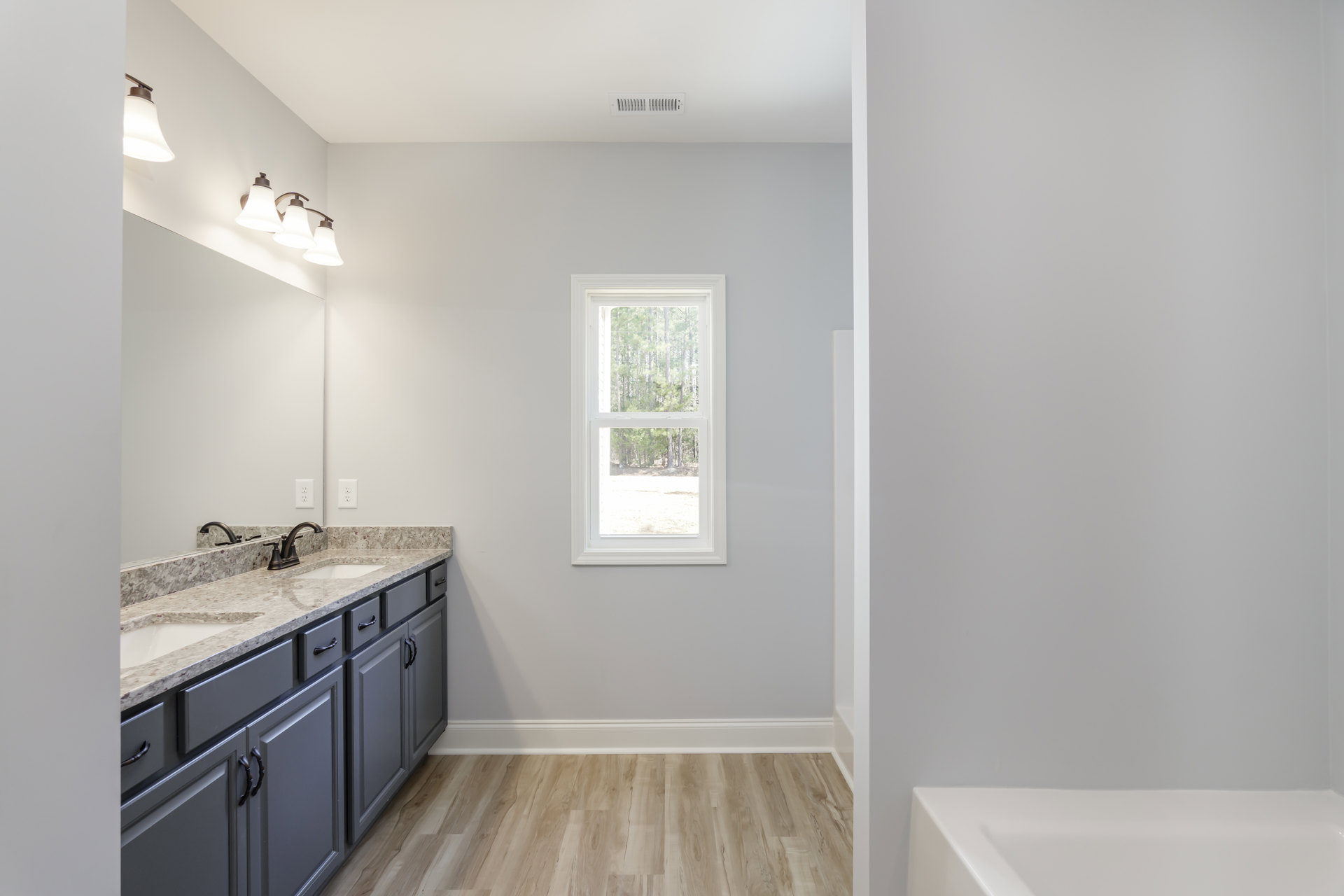 Bathroom with wood floor and white trim, white countertop and sink, chrome faucet, window overlooking trees, light fixture mounted above sink, white tile backsplash, and cabinetry