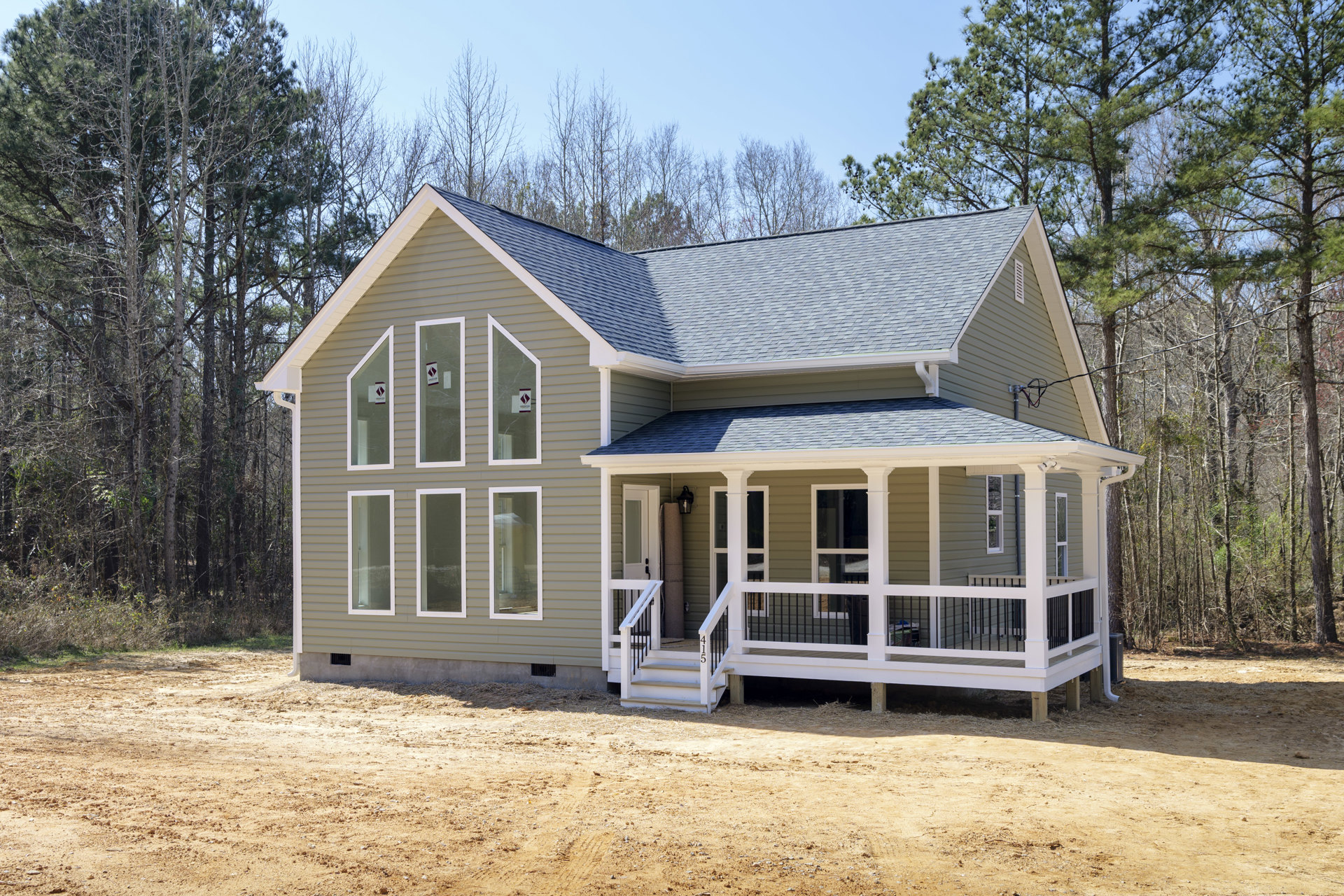 Two-story cottage-style home with white porch railing, glass door with sign, white-framed windows, and dirt path leading to entrance, surrounded by trees.