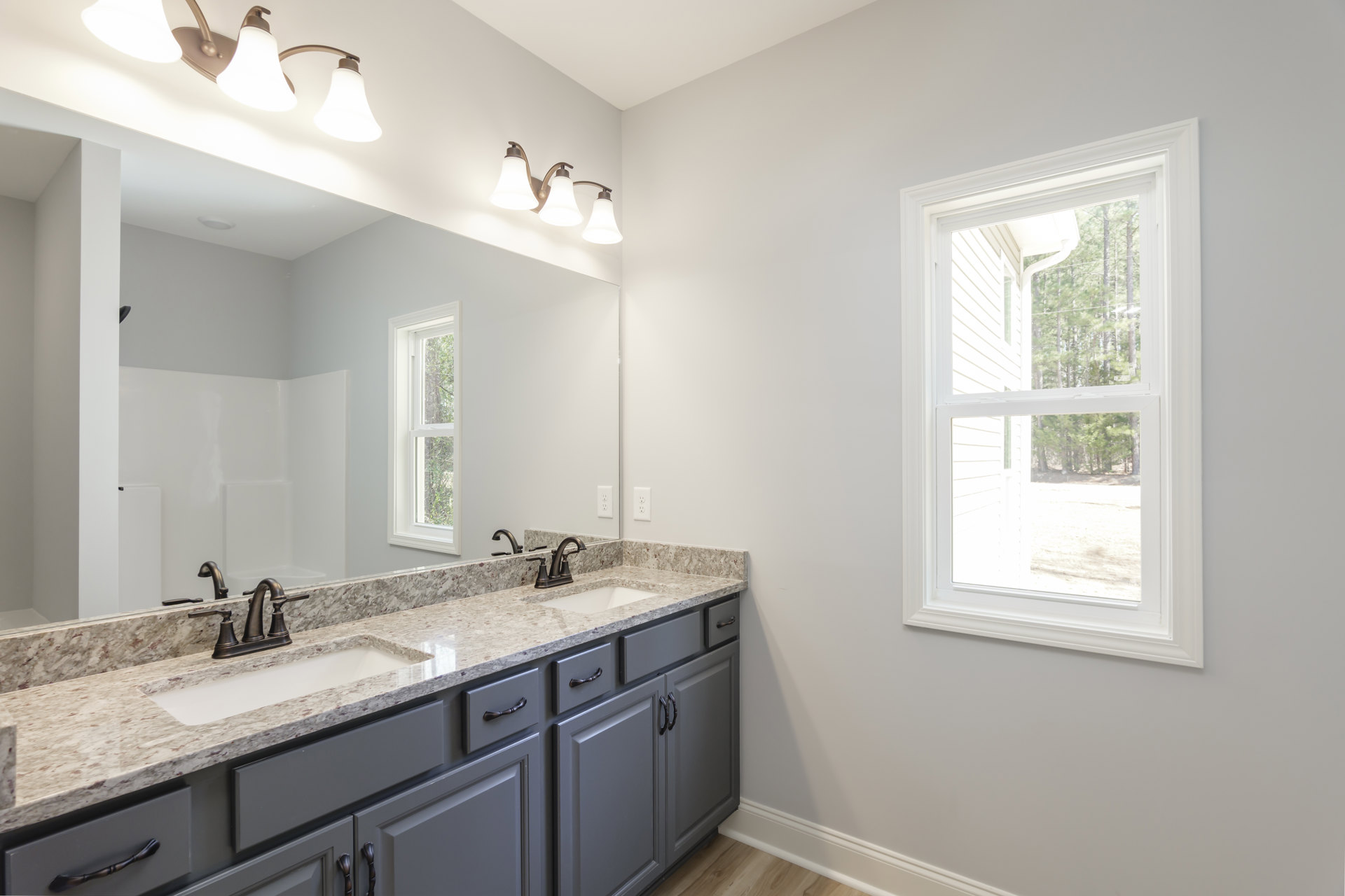 Bathroom with marble countertop and double sinks, large wall mirror above, white cabinetry, chrome faucets, three-light fixture, window with view of trees, tiled walls.