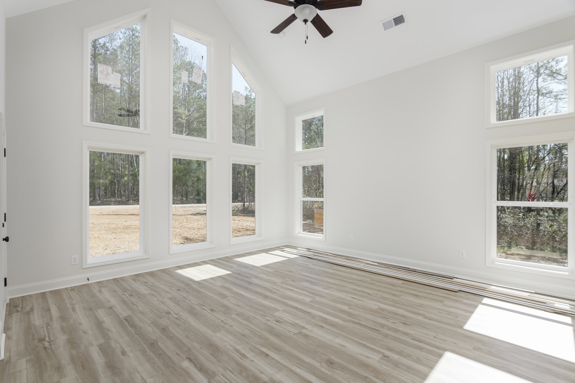 Hardwood floor room with large windows, ceiling fan with light fixture, white walls, ceiling vent, paper taped to window pane