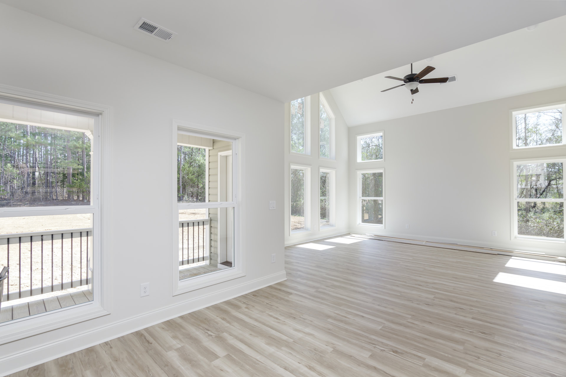 Living room with wood flooring, ceiling fan with light fixture, large windows overlooking trees, and glass door opening to balcony with metal railing