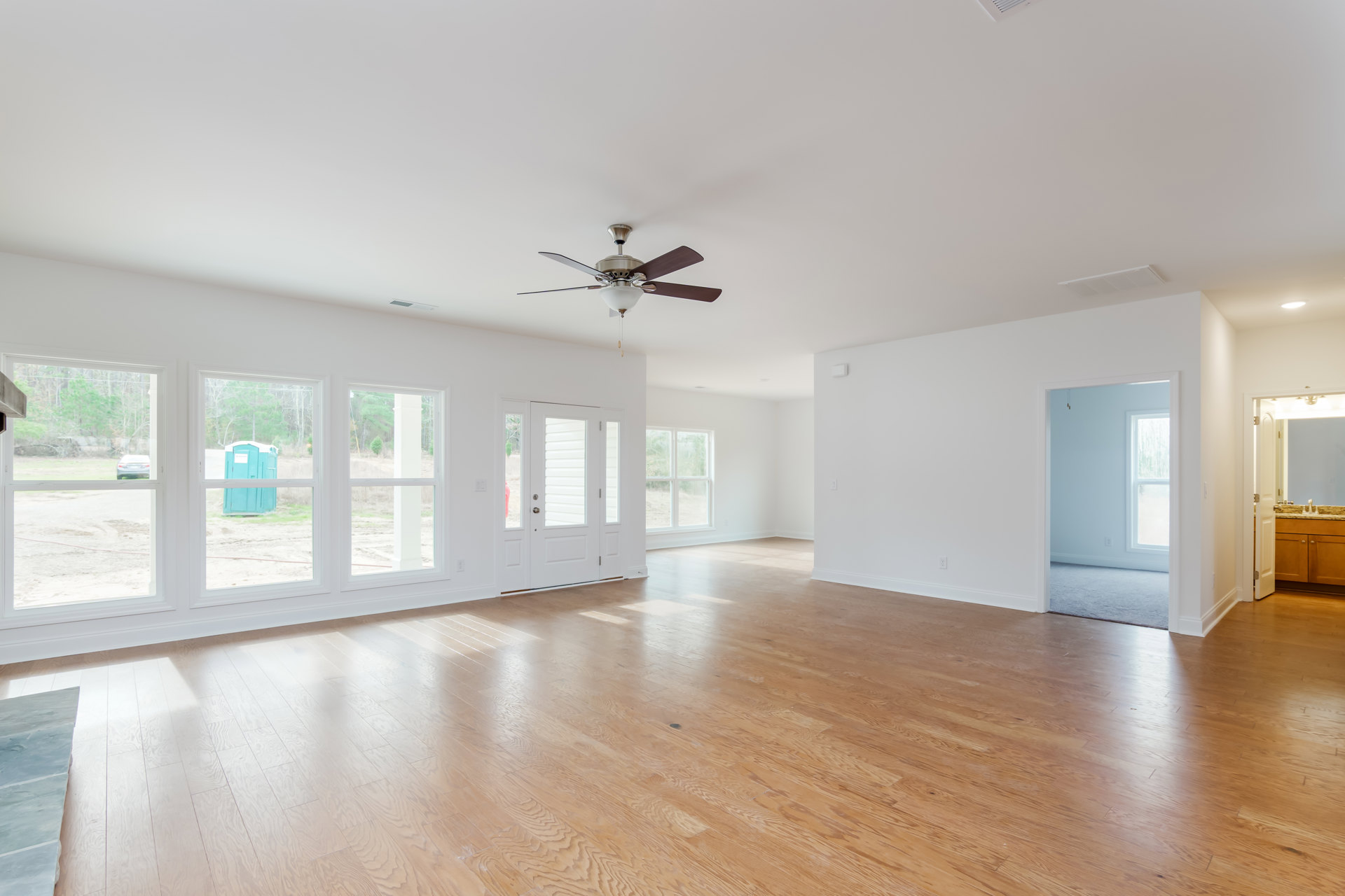Wood flooring with white doors, ceiling fan and light fixture, windows along one wall, neutral painted walls, construction visible outside through window including blue portable