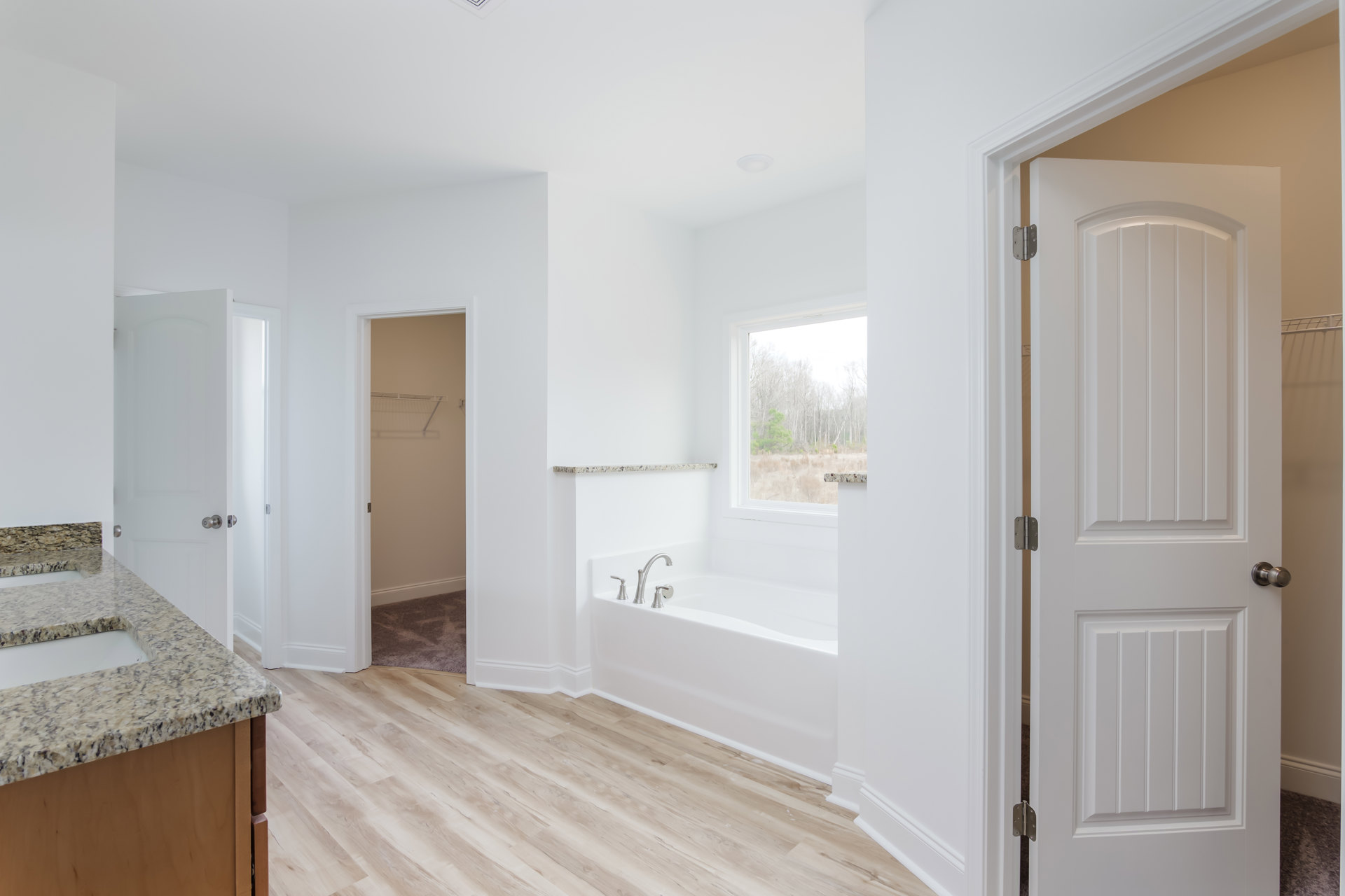 Bathroom with freestanding bathtub, wood flooring, marble countertop vanity, white walls, chrome faucet, window overlooking forest, and paneled door with metal doorknob