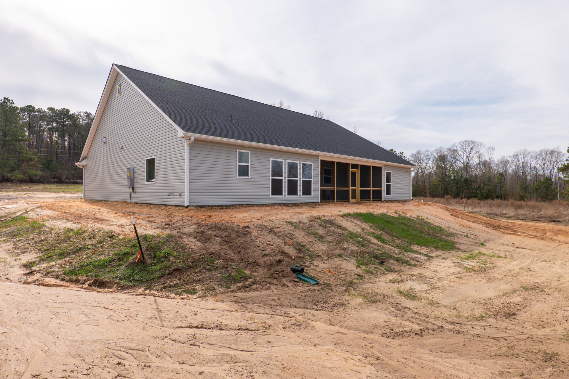 Tan dirt hill with a black pipe and drain in foreground, shovel resting on slope, two-story house in background featuring row of windows, gray roof, covered porch with door and