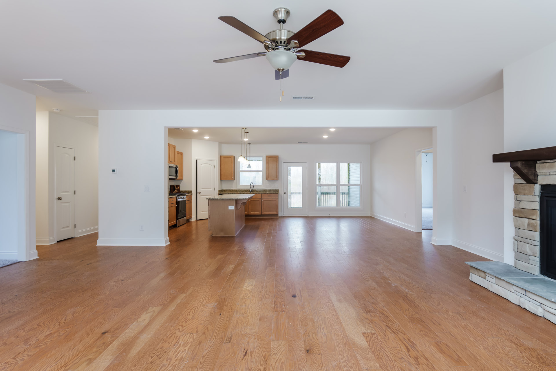 Open-concept living room with hardwood flooring, ceiling fan with light fixture, white walls, and adjacent kitchen area with cabinetry and door.