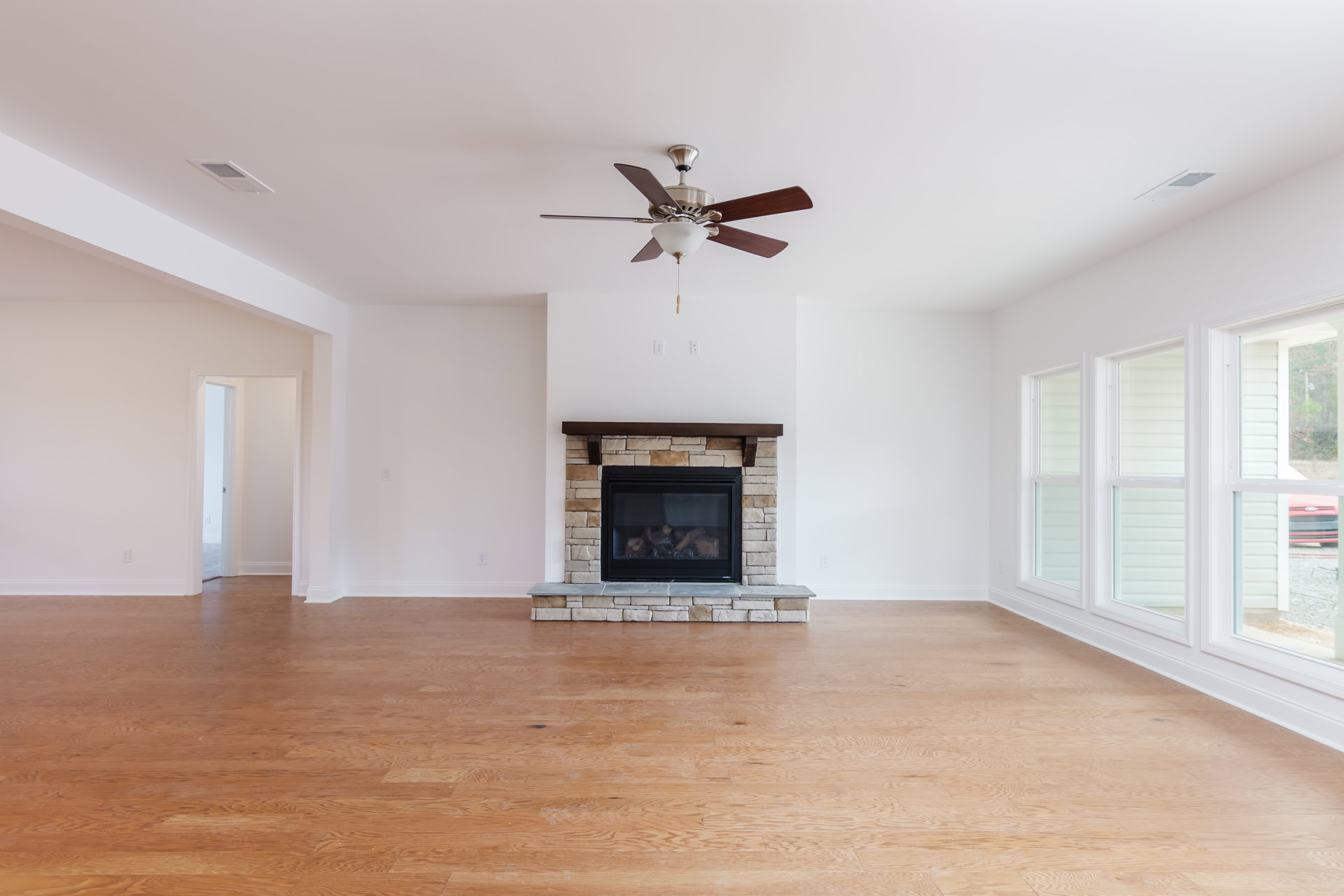 Living room with stone fireplace burning wood, wood flooring, ceiling fan with light fixture, and neutral walls.