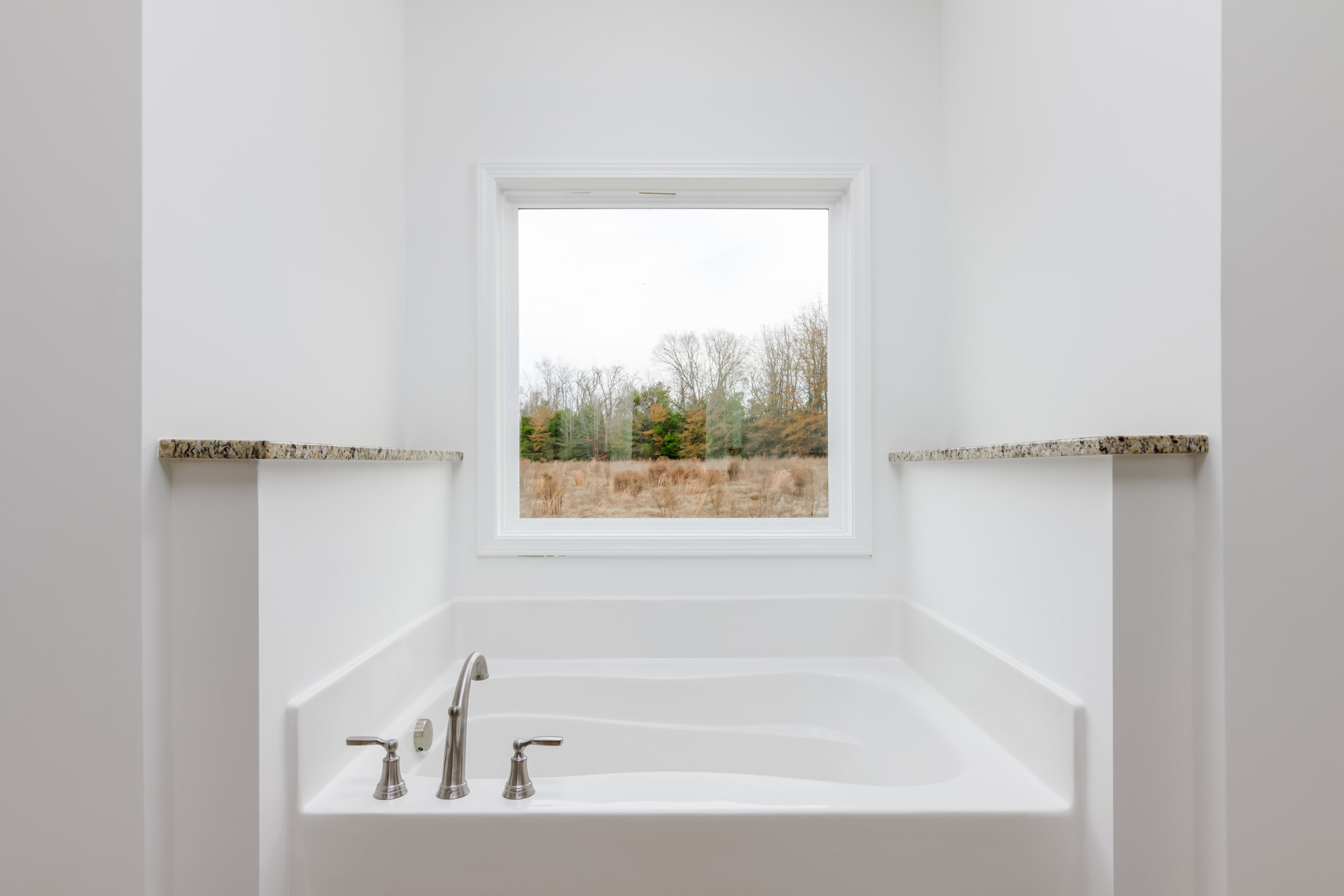 Freestanding white bathtub beneath a large window with tree views, chrome faucet mounted on tub, light-colored bathroom walls, natural light illuminating space