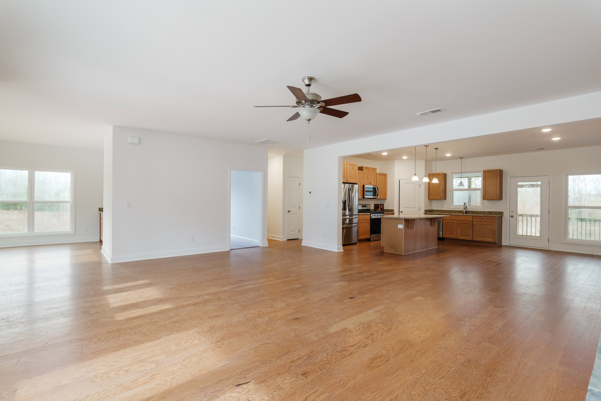 Spacious open floor plan featuring wood flooring, white walls, modern kitchen with stainless refrigerator, glass panel door, multiple windows, and ceiling fan.
