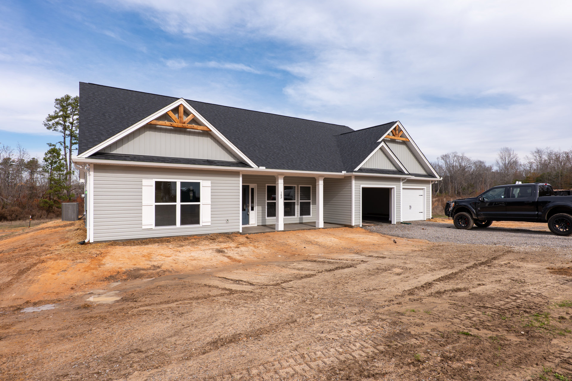 Modern home with grey roof, white siding, and large windows, black pickup truck parked on gravel driveway, attached garage, metal utility box near front yard, trees and cloudy sky