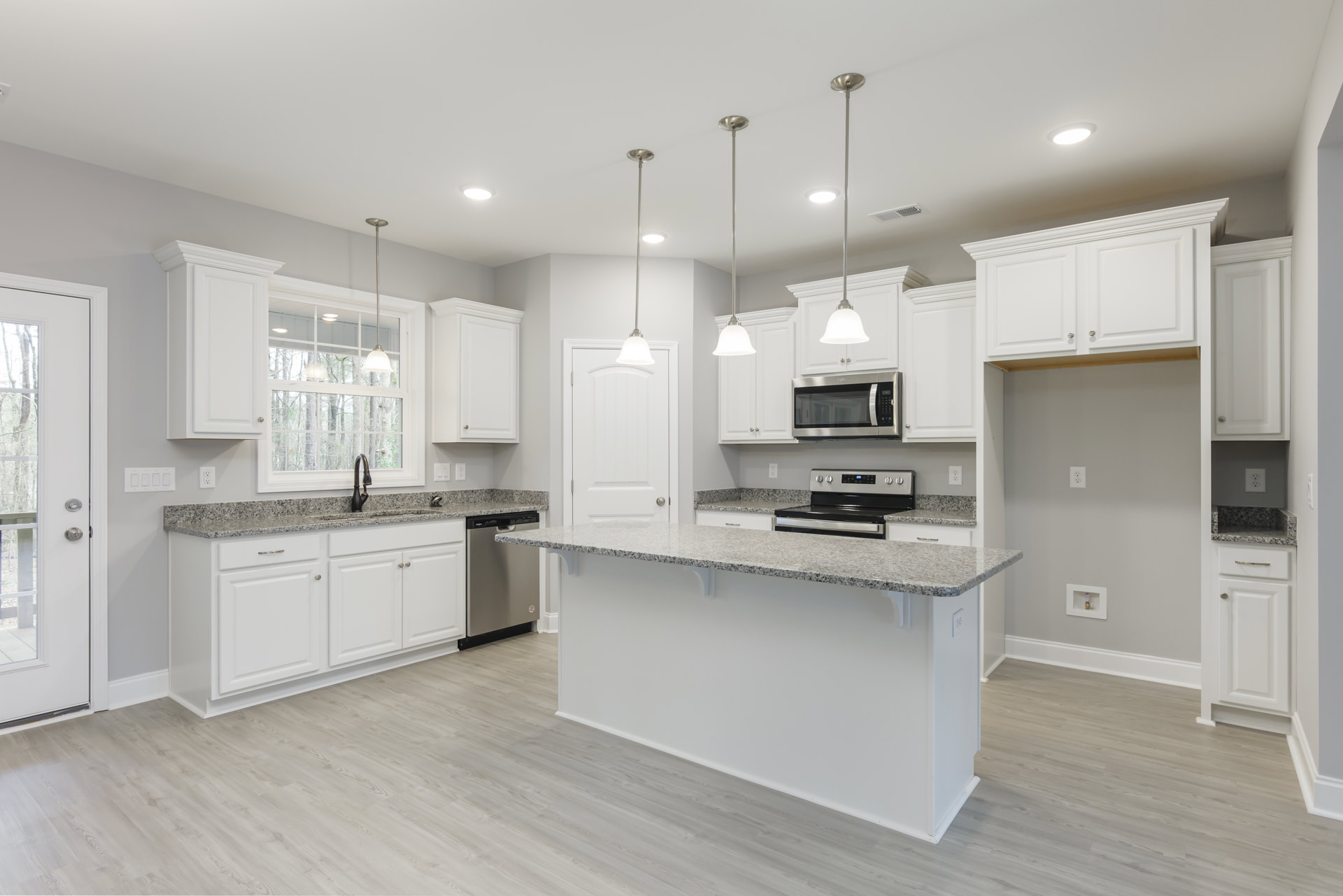 White kitchen with granite countertops, stainless steel oven and microwave, white cabinetry, ceiling light fixture, and adjacent white door with silver knob.