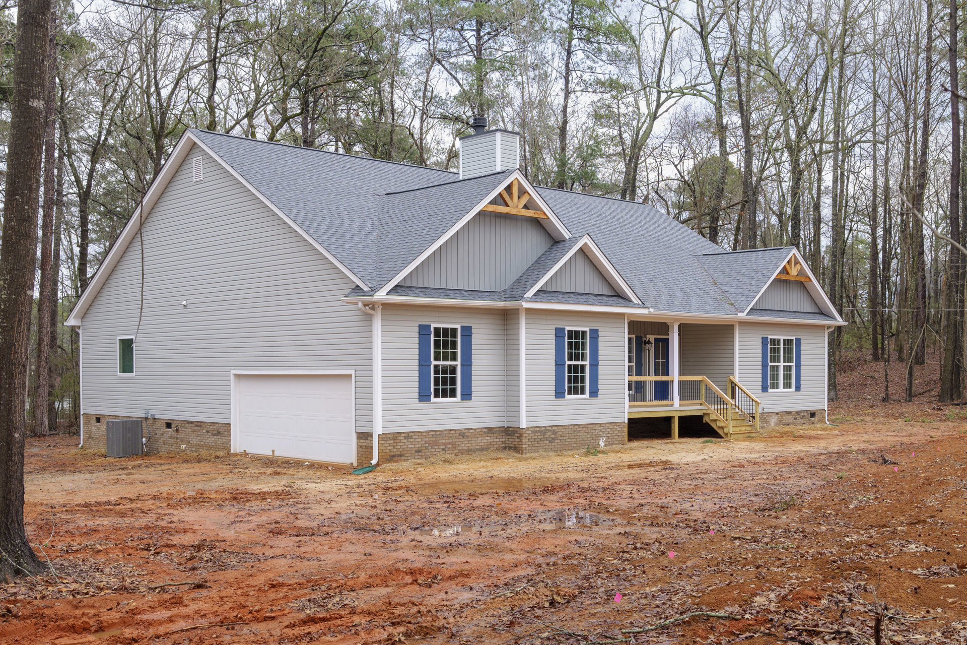 Partially built house with exposed wooden deck and railing, white garage door, white roof, grey air conditioner unit beside brick siding, surrounded by trees and dirt road