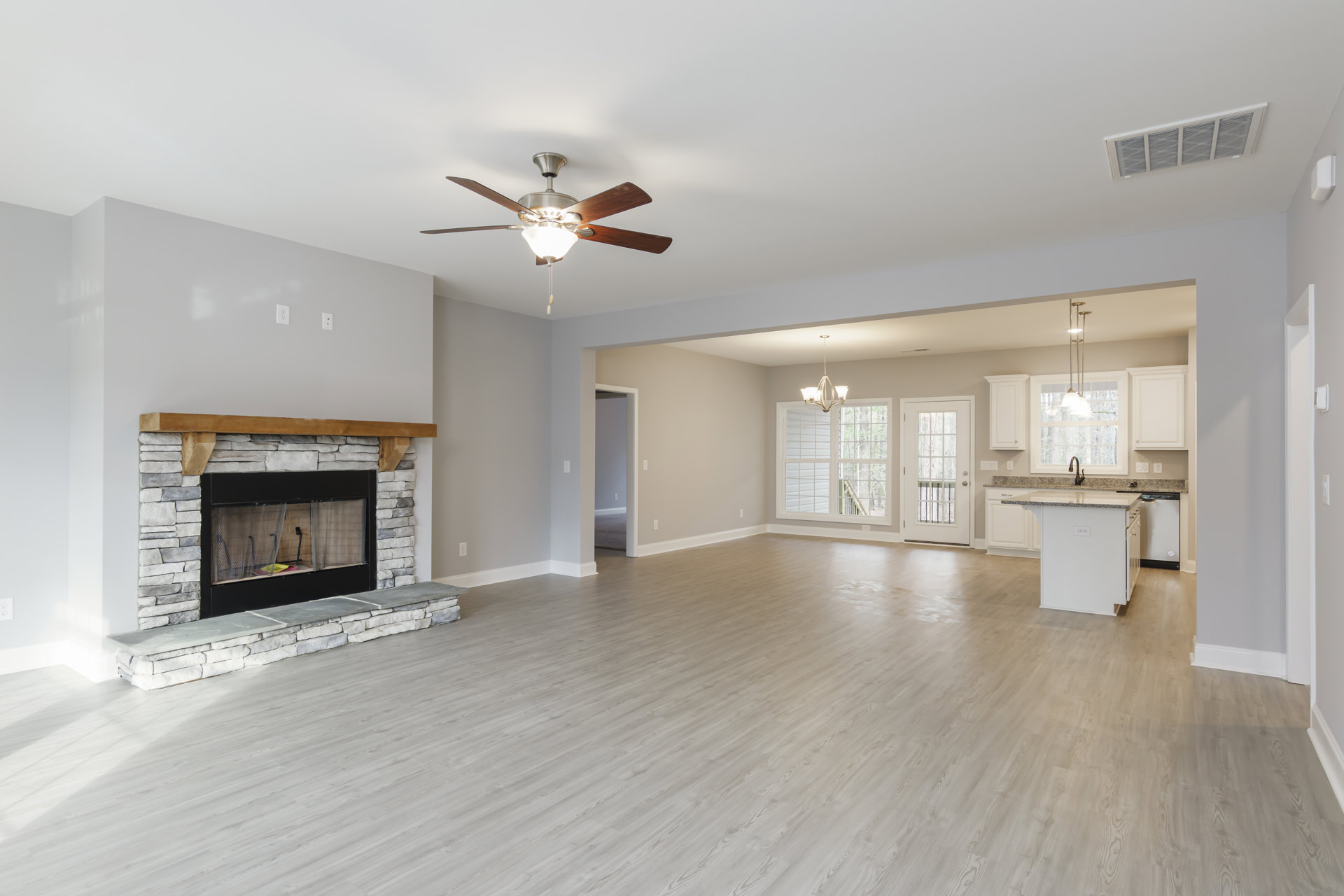Living room with wood flooring, ceiling fan with light, fireplace featuring a wood frame and screen, adjoining kitchen, white door with glass panes.