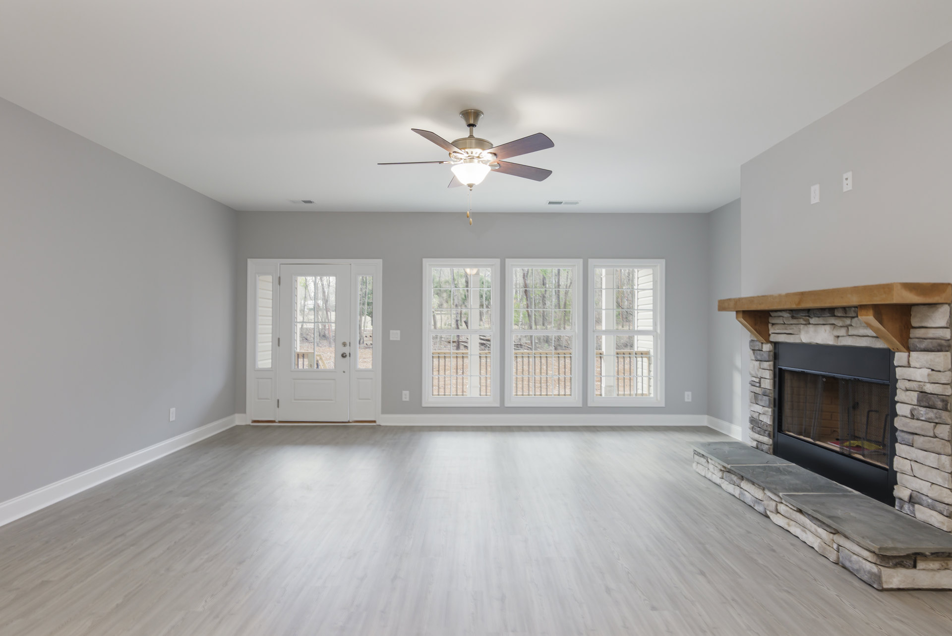 Living room with white wood flooring, white walls, ceiling fan with light fixture, fireplace featuring a wood mantel, window showing trees outside, and white door with glass panes