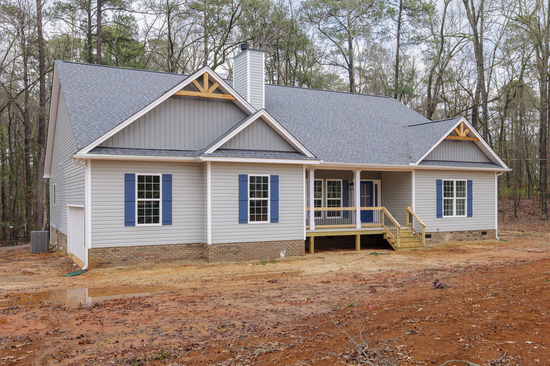 Two-story cottage with blue shuttered windows, white trim, wood siding, covered porch, wooden deck with railing, surrounded by trees, dirt road in foreground