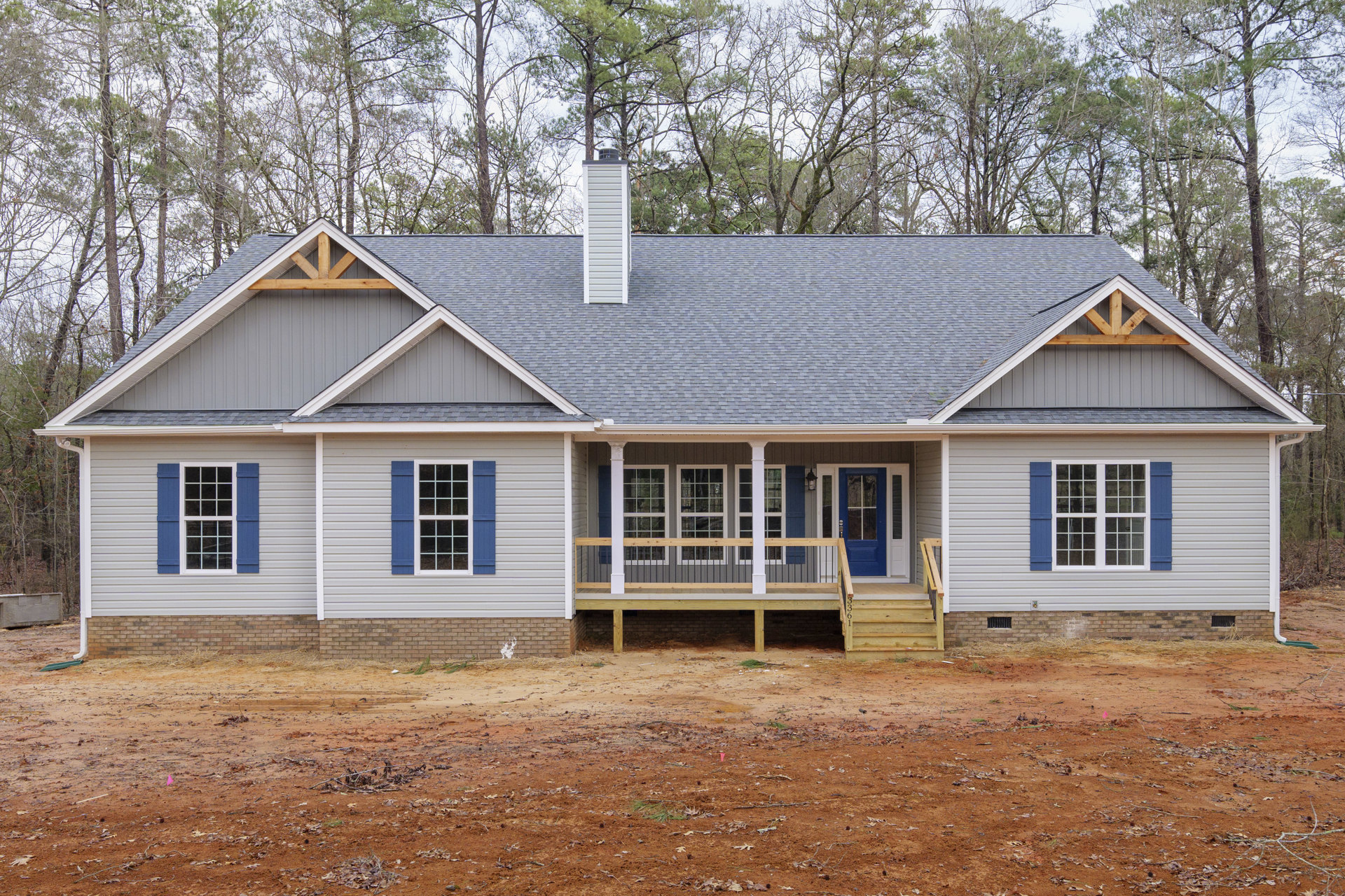 Two-story house with beige siding, blue shutters, white trim, covered porch with wooden railing, chimney, wooden steps leading to blue front door, dirt yard, and mature tree.