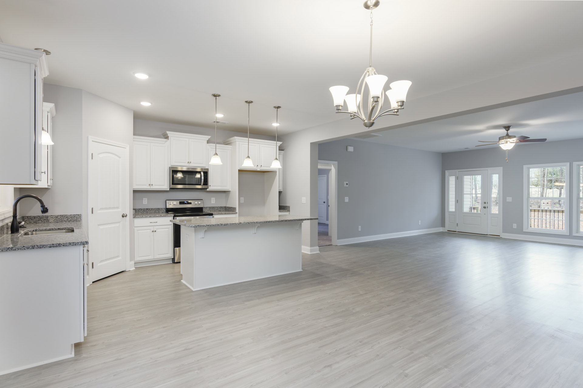 Open-concept kitchen and living room featuring white cabinetry, wood flooring, stainless steel kitchen hood, modern pendant lamp, and large window overlooking trees