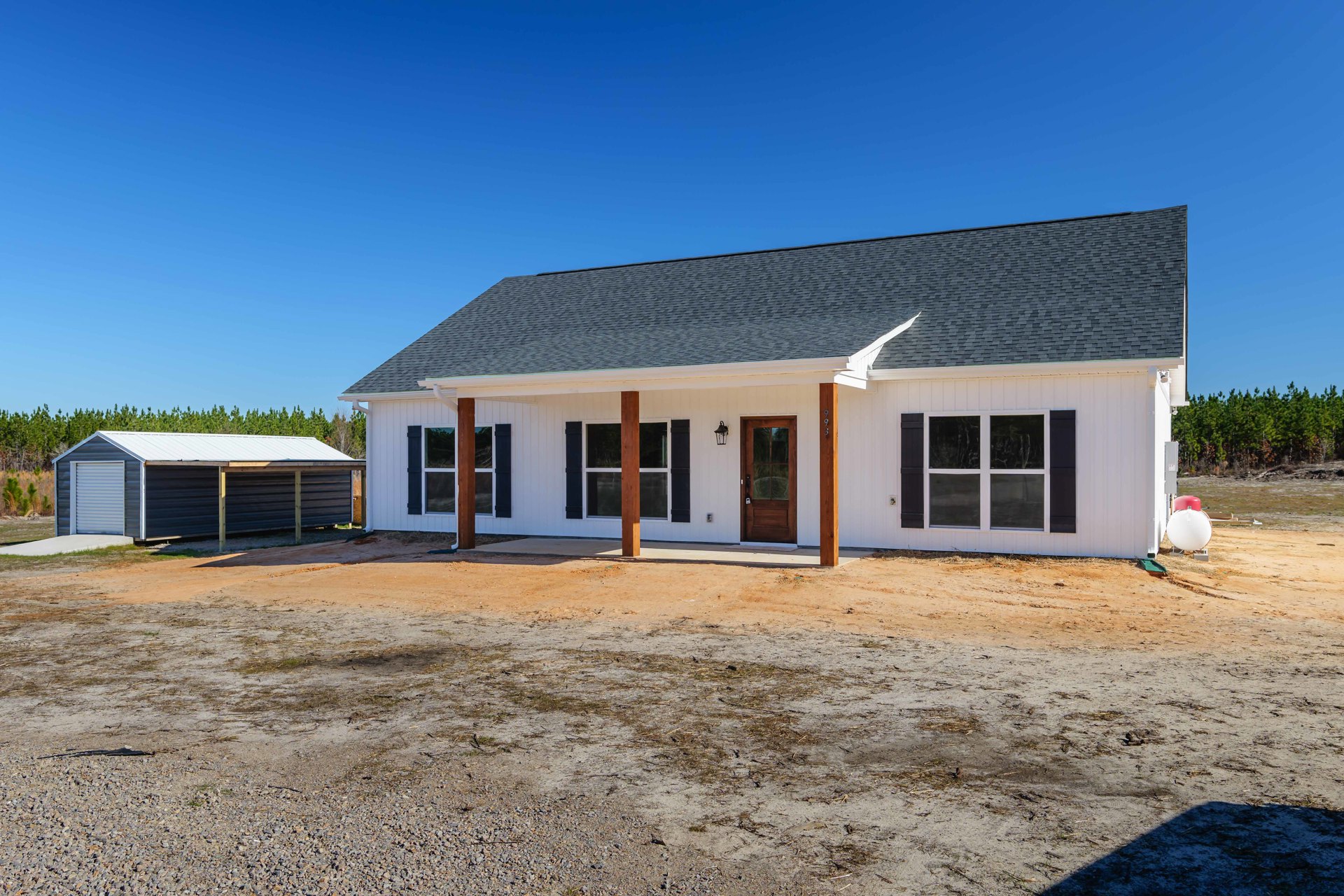 White house with black roof, dirt yard, paved driveway, shed with sloped roof, front door with key lock, windows, trees in background, white round object with red cap near