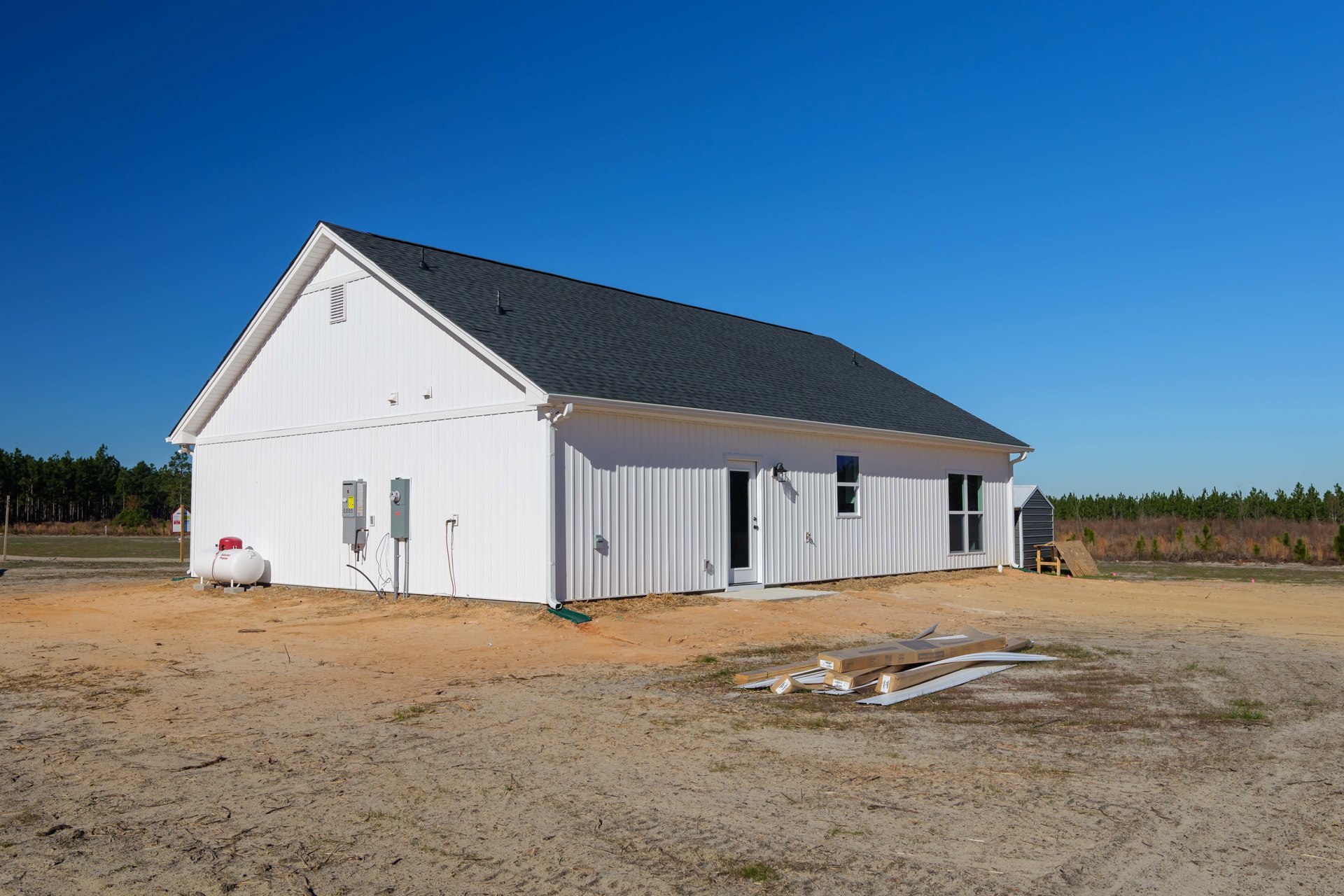 White house with black metal roof, wood planks stacked beside exterior wall, white cylindrical tank with red top, cardboard boxes on ground, construction equipment nearby, open lot