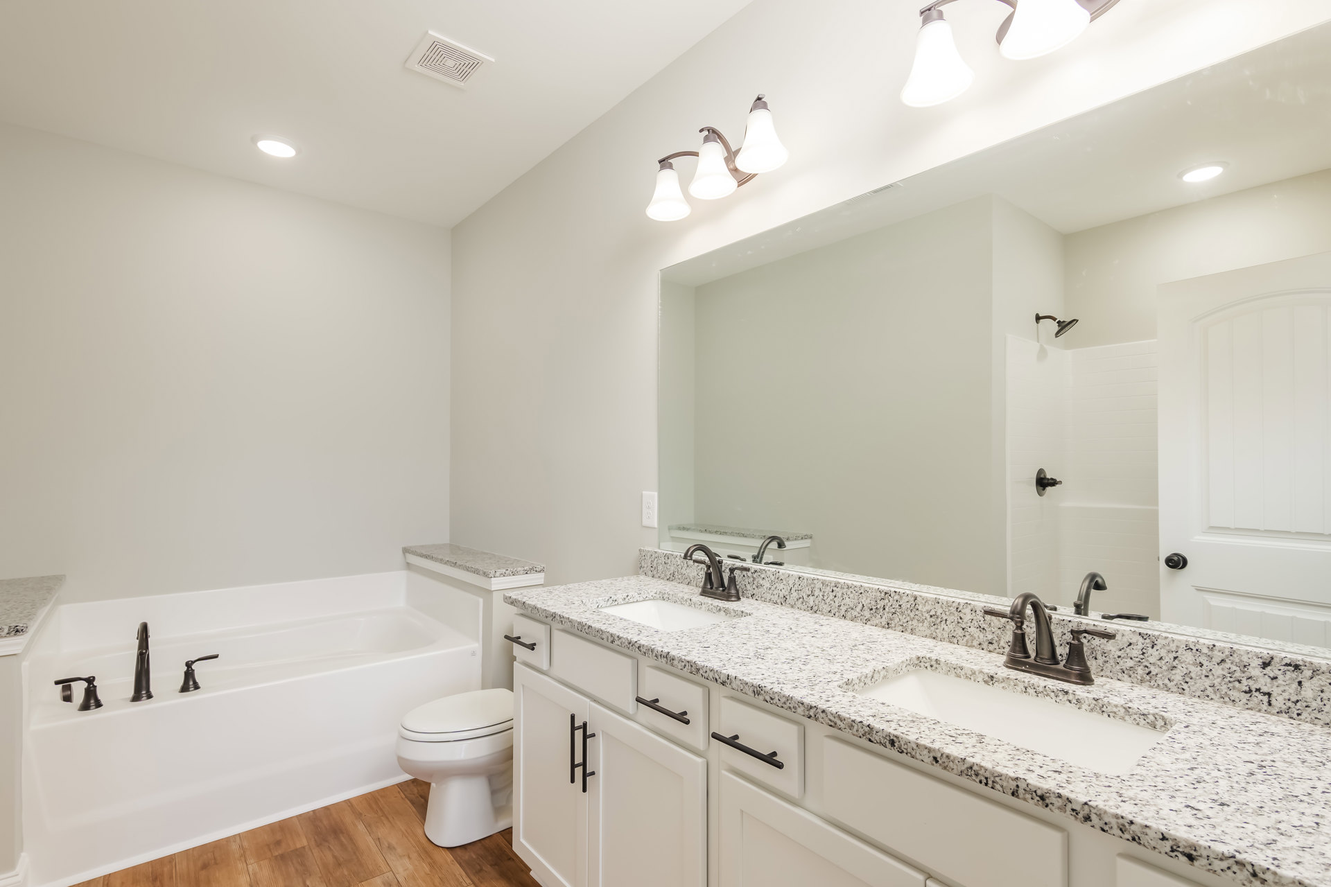 Marble countertop vanity beside a freestanding bathtub, white toilet on wooden floor, three-light ceiling fixture, vent, and close-up of light bulb