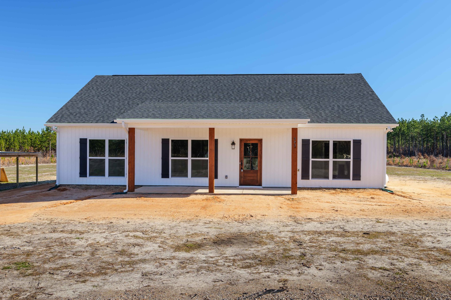 White siding home with brown shingle roof, brown front door, multiple windows, dirt yard, and sparse landscaping under blue sky.