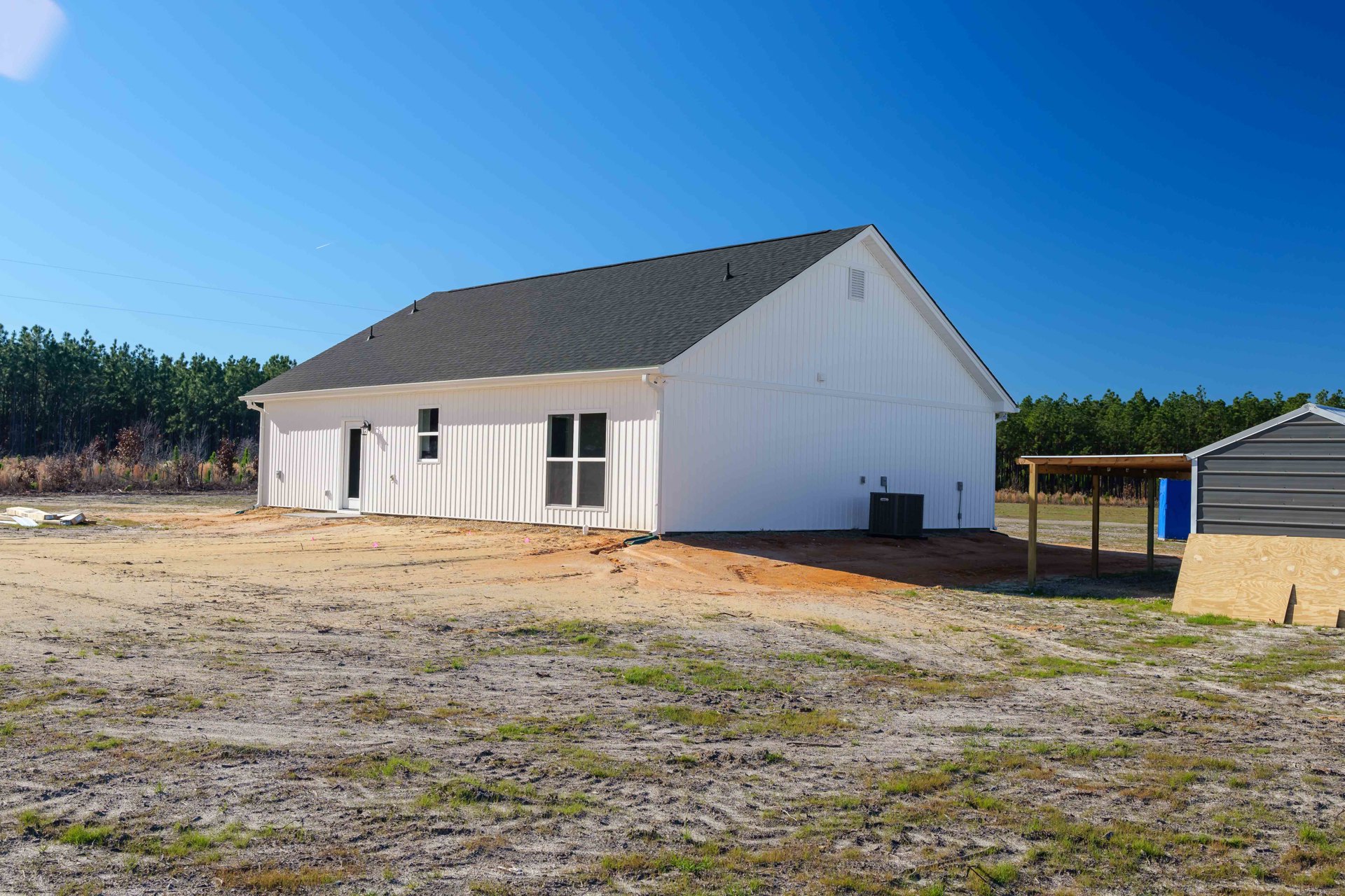 White house with black roof and central shed, surrounded by dirt field with patches of grass; plywood sheets lie near a road, visible window and roof vent.