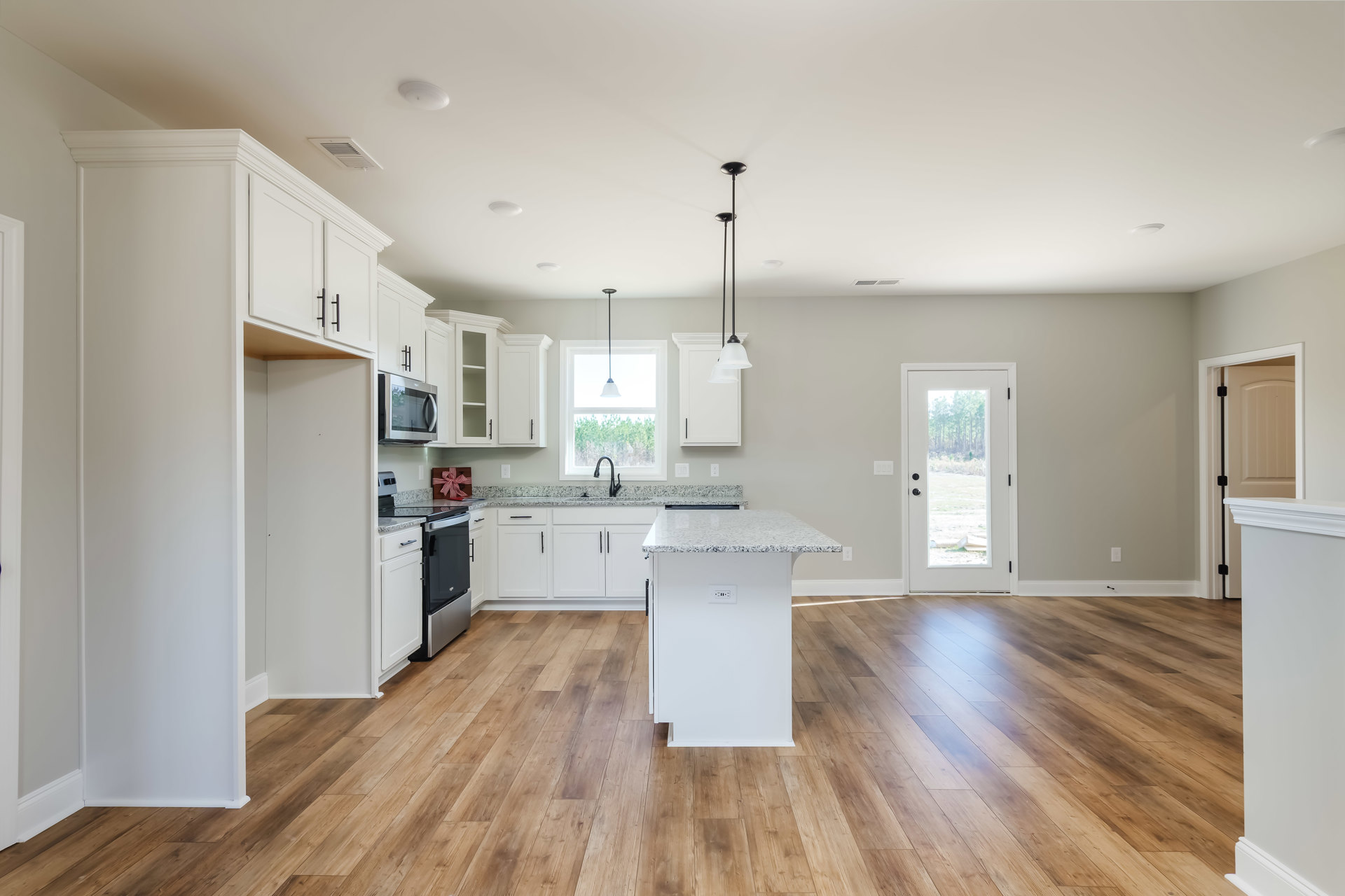 White kitchen with wood flooring, white cabinets, marble-topped island, open microwave door, door with window showing trees, gift wrapped in red and white ribbon on countertop
