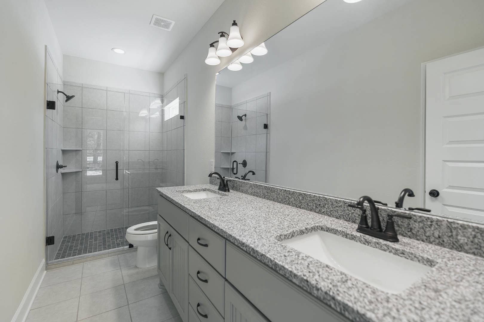 Bathroom with a wide vanity mirror, white sink, row of modern light fixtures, tiled walls, and a rectangular window letting in natural light.
