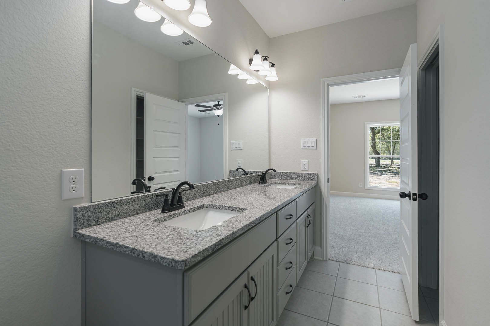 Bathroom with double sinks, wide mirror above, white countertop, black-edged sinks, tile flooring, cabinetry, window showing leafy tree outside, white electrical outlet, carpeted