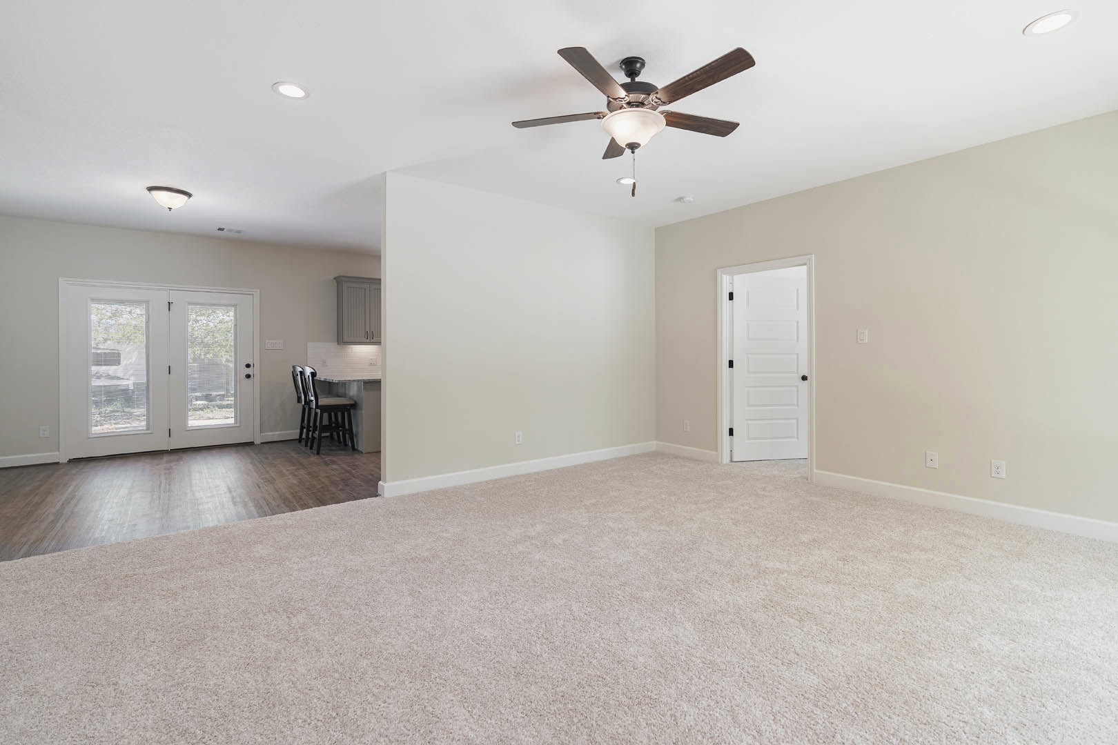 Open-concept living area with laminate flooring, ceiling fan with light fixture, carpeted section, white walls, double glass-paneled doors, white door with black knobs, and kitchen