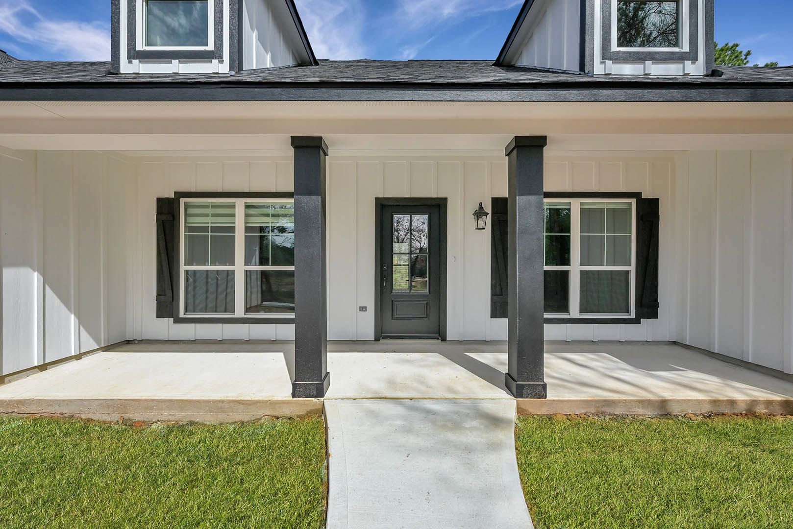Modern home with gray front door, concrete walkway, covered porch, large windows, manicured grass lawn, and blue sky
