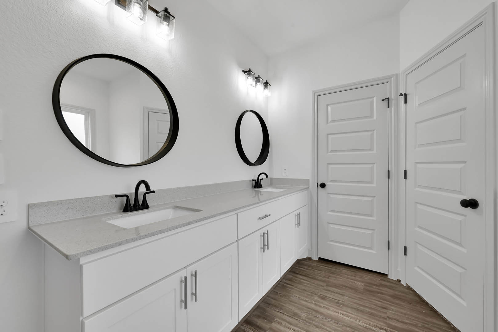 White bathroom cabinets with black round mirrors above, black faucet on white countertop, white door with black knob, light tile flooring.
