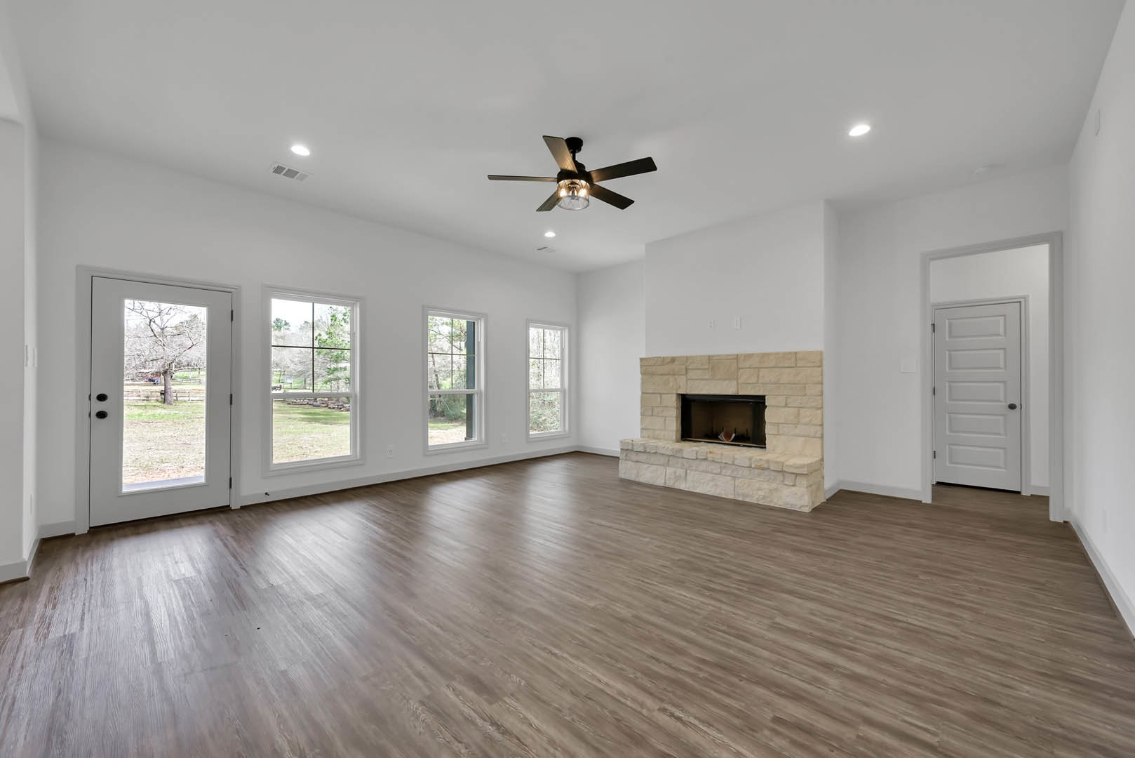 Living room with wood flooring, white walls, ceiling fan with light, fireplace with white mantel, and white door with black knob leading outside to a tree.