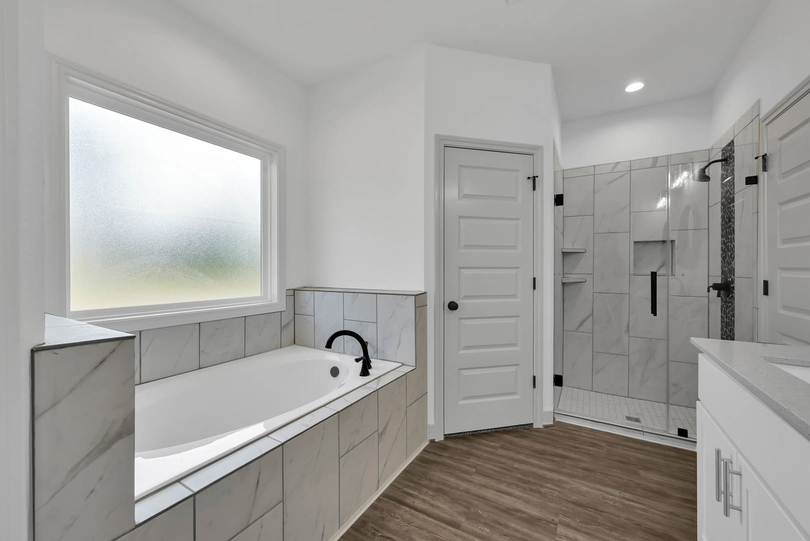 Bathroom with freestanding white bathtub and black faucet, glass-enclosed shower, frosted window, wood flooring, white door with black knob, white sink with black faucet, light