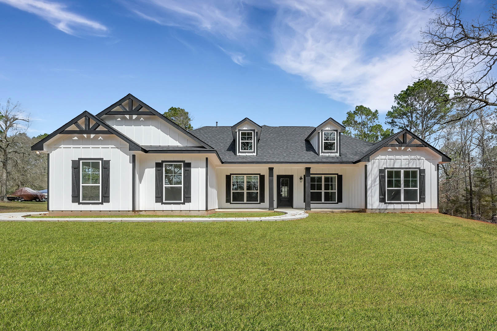 Two-story house with black roof, white-framed windows, and covered porch, surrounded by green lawn; leafless tree and covered boat on trailer in front yard