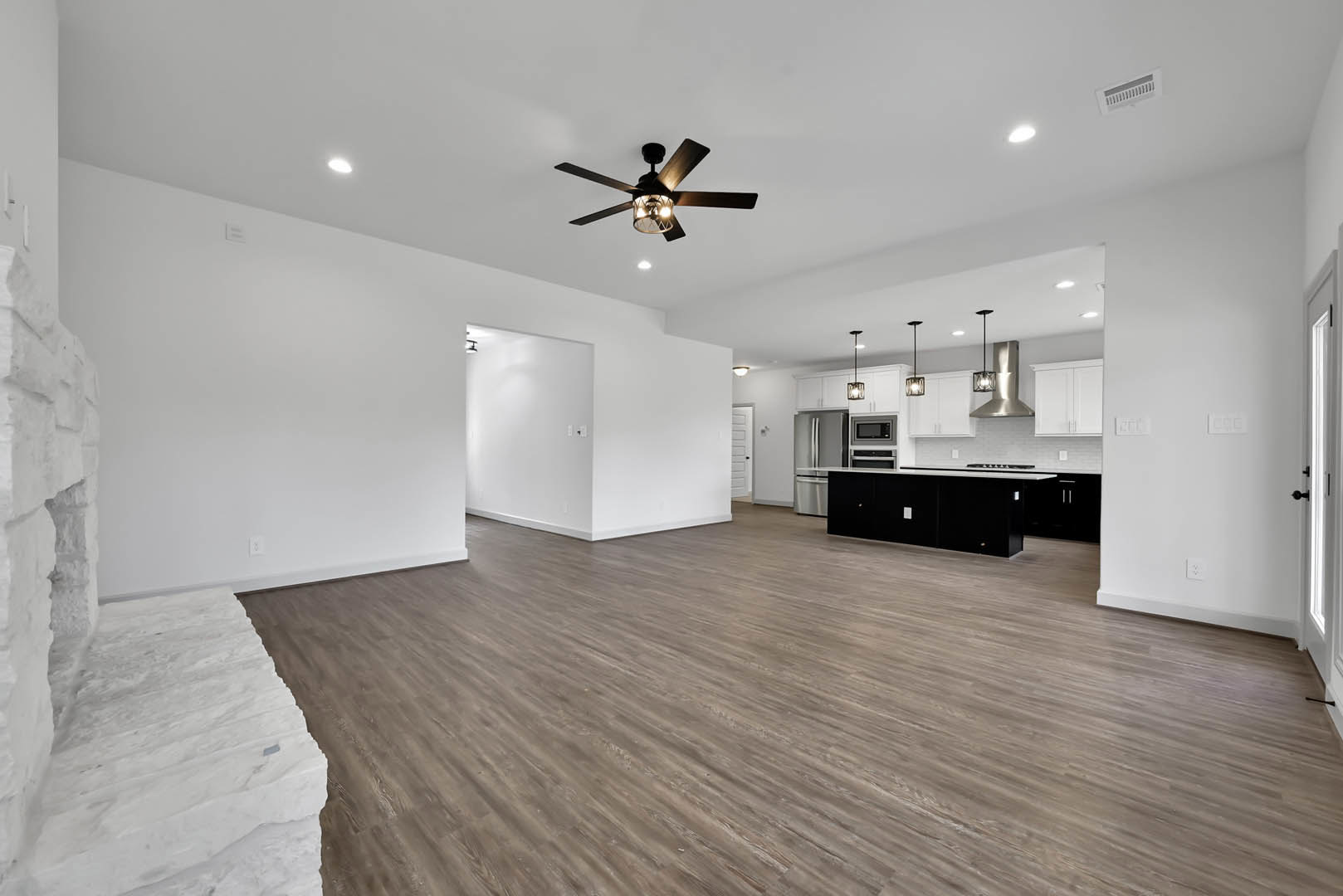 Open-concept room featuring wood flooring, white stone accent wall, ceiling fan with light, wall vent, and partial view of a stainless steel refrigerator in the adjacent kitchen