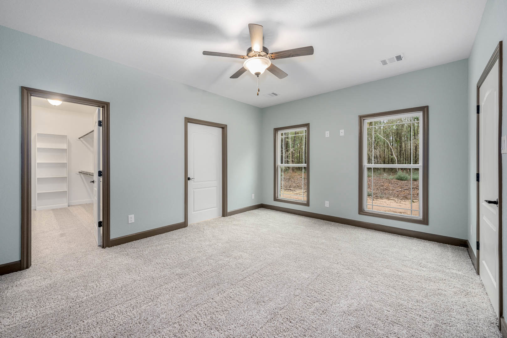 Carpeted room with a ceiling fan and light fixture, large window overlooking forest, white shelving unit, and white door with black handle