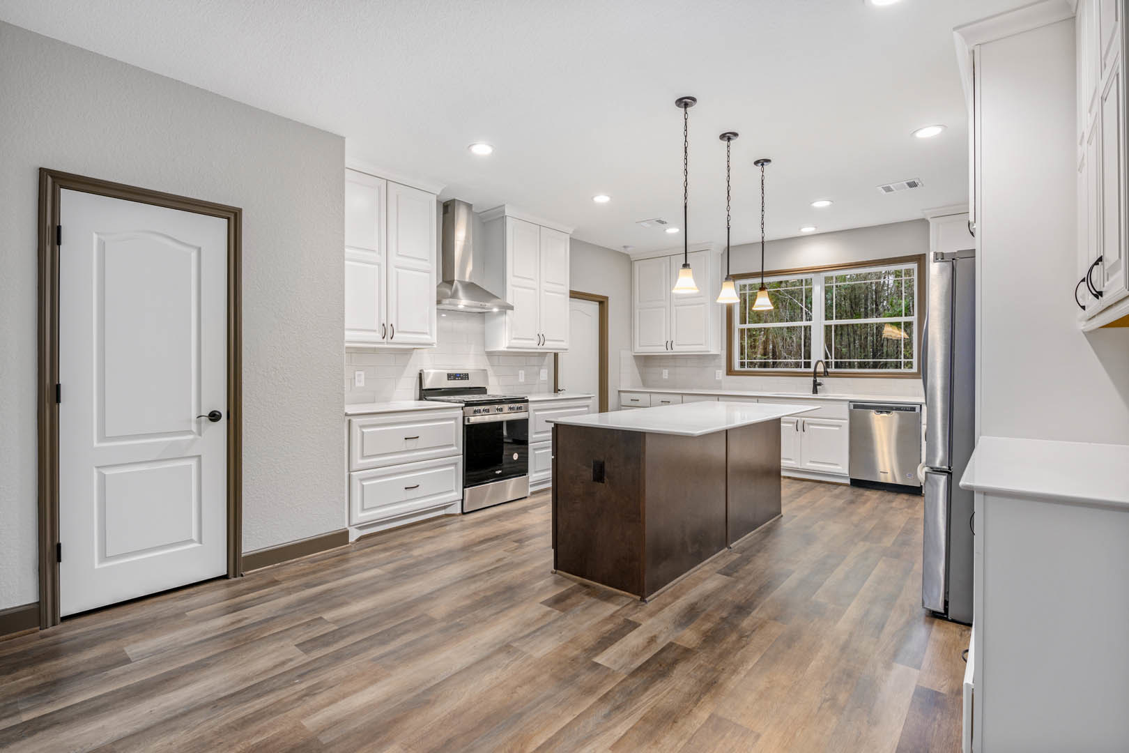 Kitchen with light wood flooring, white shaker cabinets, white quartz island countertop, stainless steel stove and oven, black hardware on a white door, large window overlooking