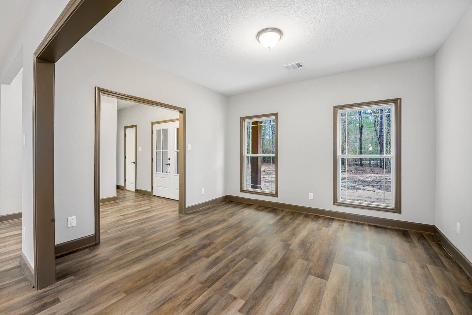 Spacious room with light wood flooring, large windows overlooking leafy trees, white door with glass panes, and modern ceiling light fixture