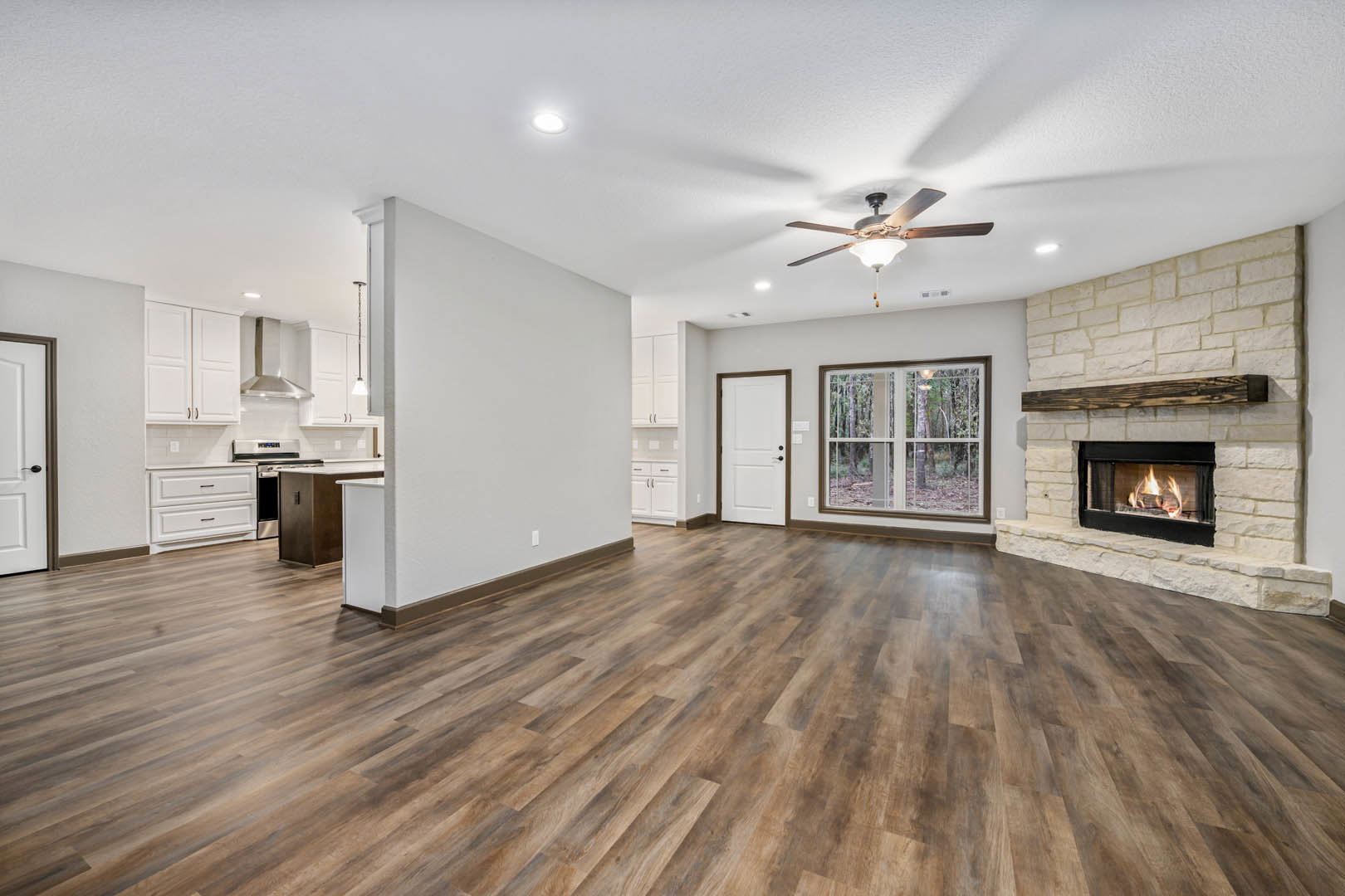 Living room with hardwood floors, plaster walls, central fireplace with burning fire, ceiling fan with light fixture, large window overlooking forest