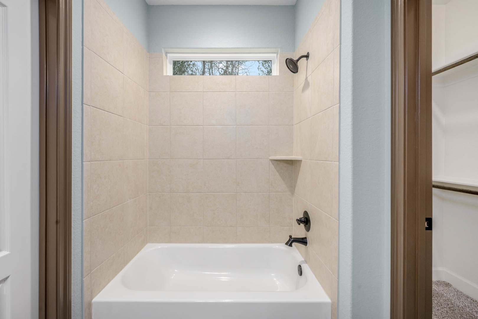 Freestanding white bathtub beside a window with white frame, wall-mounted shower head, white tile walls, chrome door knob, and bathroom accessories