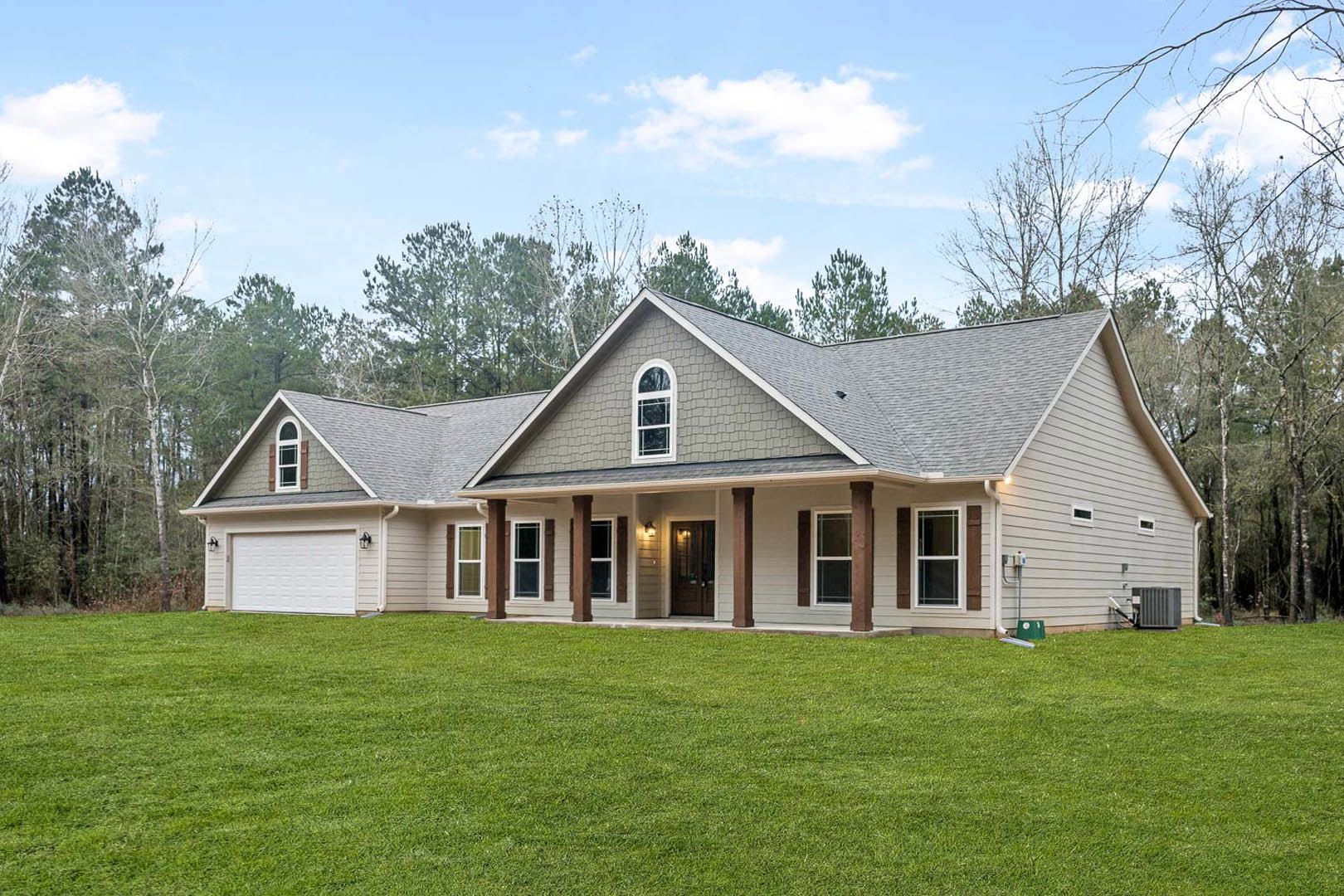 Two-story house with large covered porch, brown pillars, white-framed windows, green lawn, mature trees, and Robert Frost Farm visible in the background under a partly cloudy sky