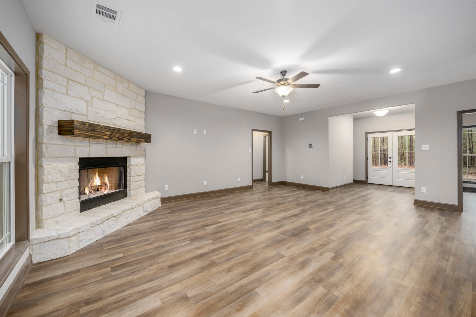 Living room with hardwood flooring, stone fireplace with burning fire, ceiling fan with light, double glass-paneled doors, wood plank accent wall, and plaster walls.
