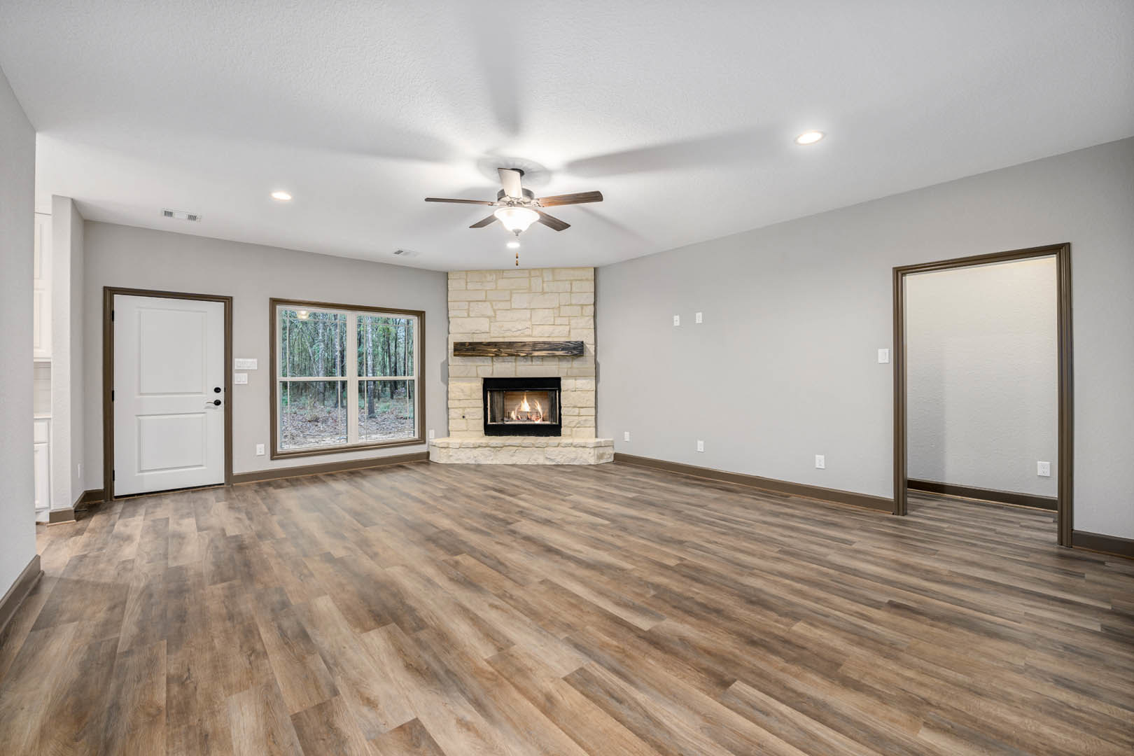 Living room with wood flooring, white walls, brown trim, ceiling fan, fireplace with burning fire, large window showing trees outside, white door with black handle