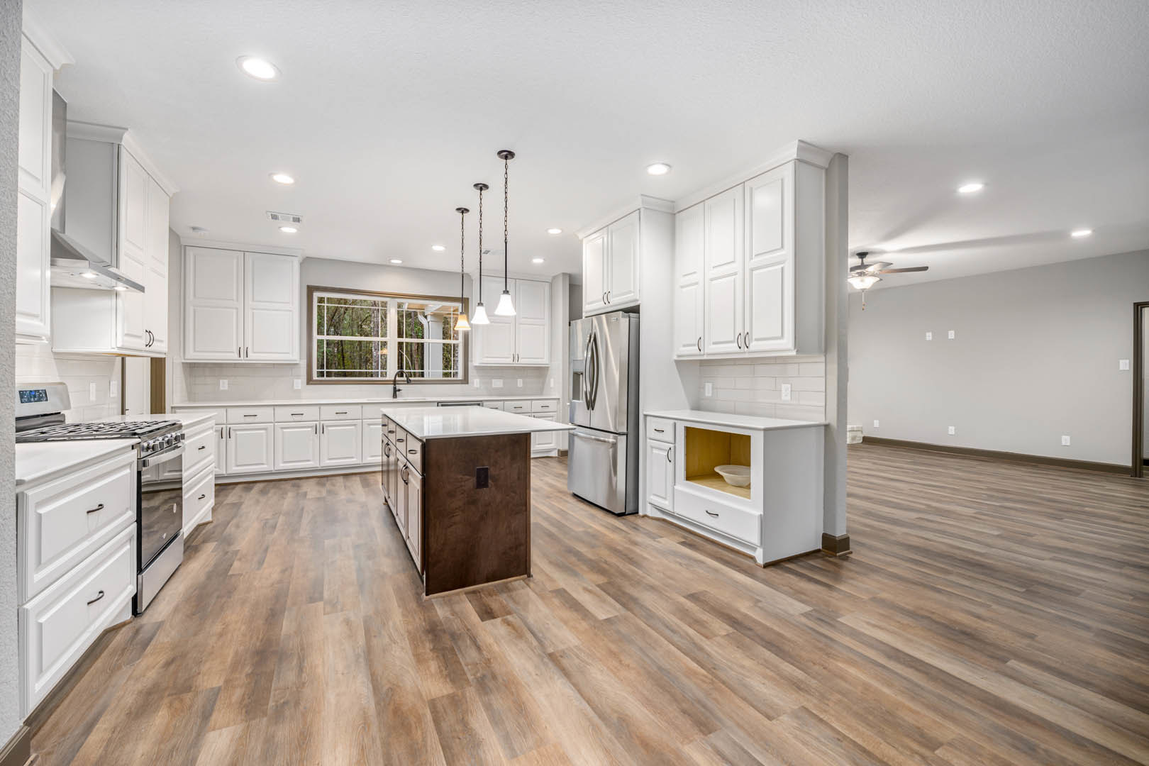 White kitchen cabinets, wood flooring, kitchen island with white countertop, stainless refrigerator, window showing trees, open cabinet with bowl inside