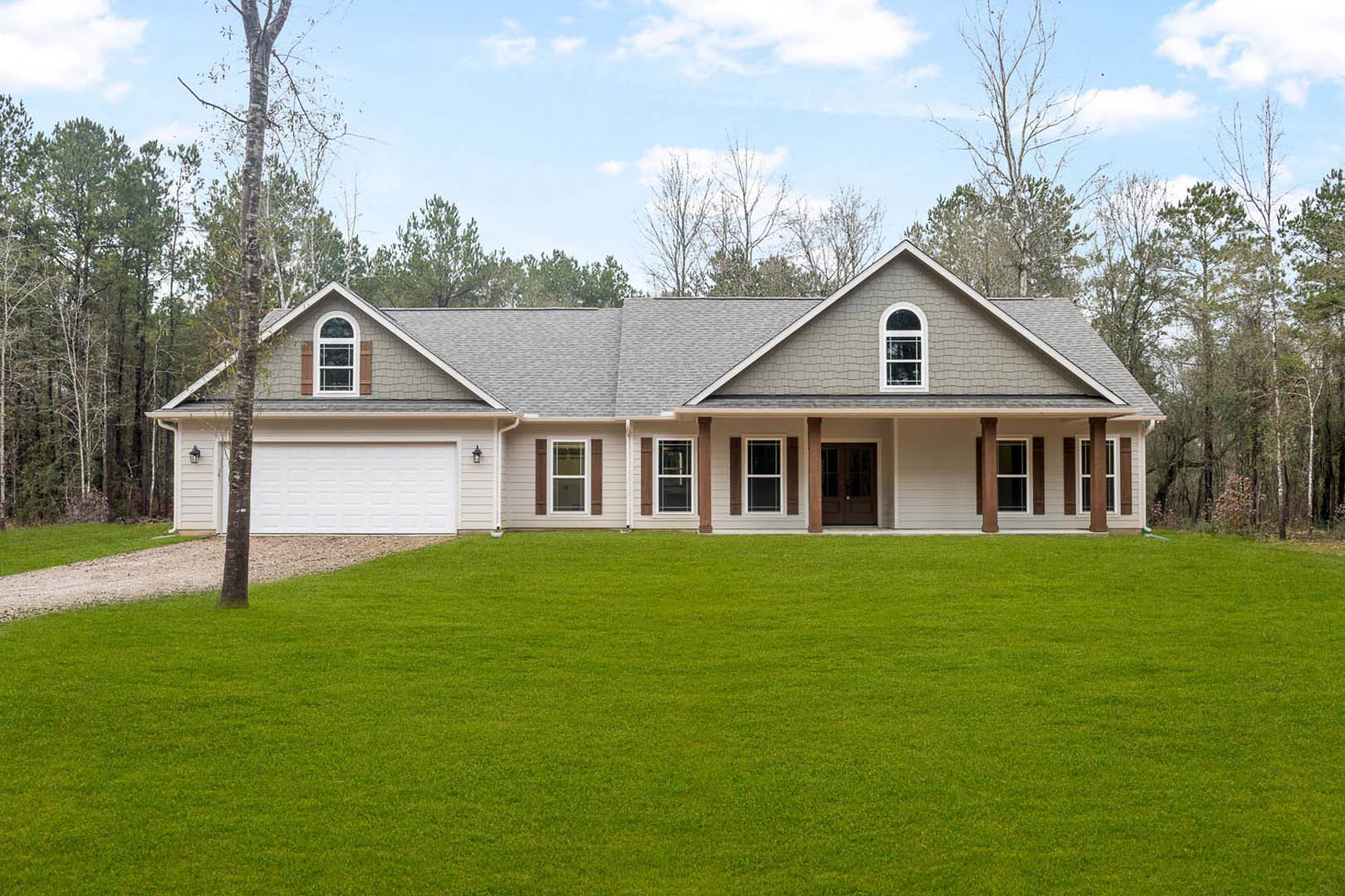White two-story house with large windows, attached garage, green lawn, mature trees, and covered front porch under a partly cloudy sky