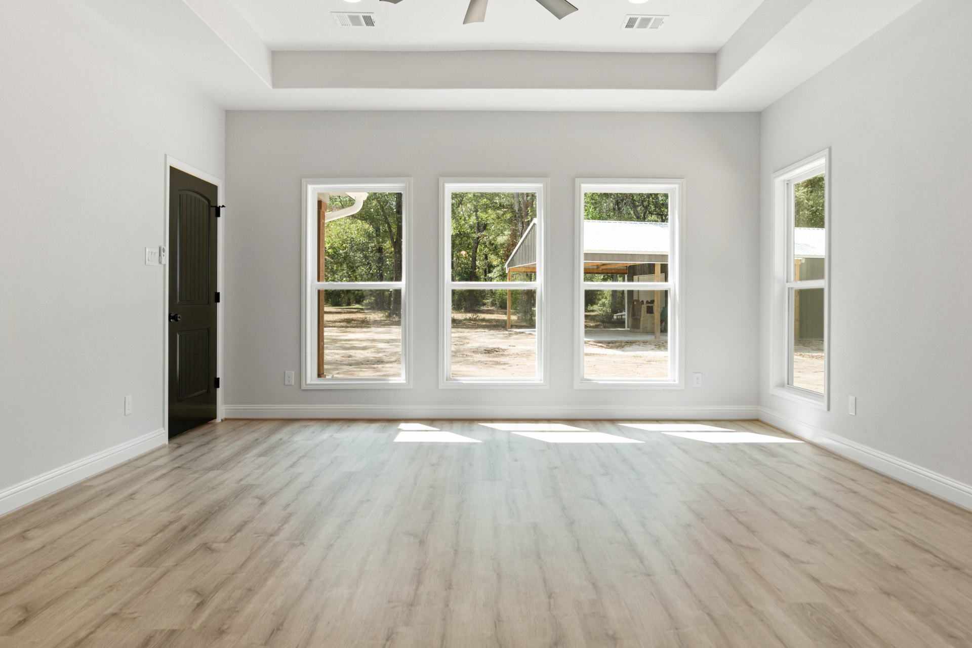 Wood flooring with sunlight streaming in, black door, ceiling fan, large windows framing leafy trees, white walls, and plaster finishes.
